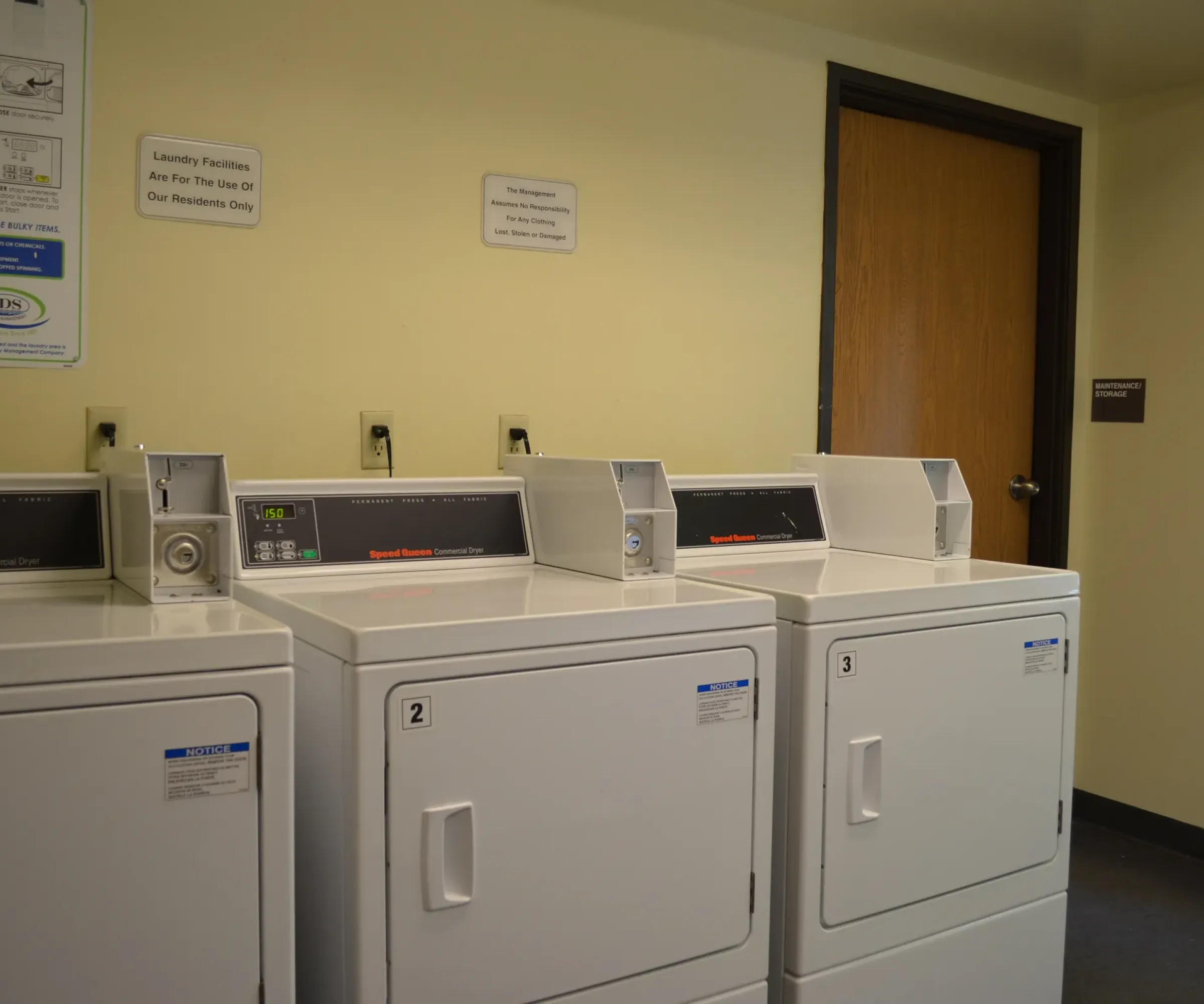 Aeon Affordable Homes and Community Laundry room in affordable apartment homes, featuring three white washing machines against a beige wall by a wooden door.