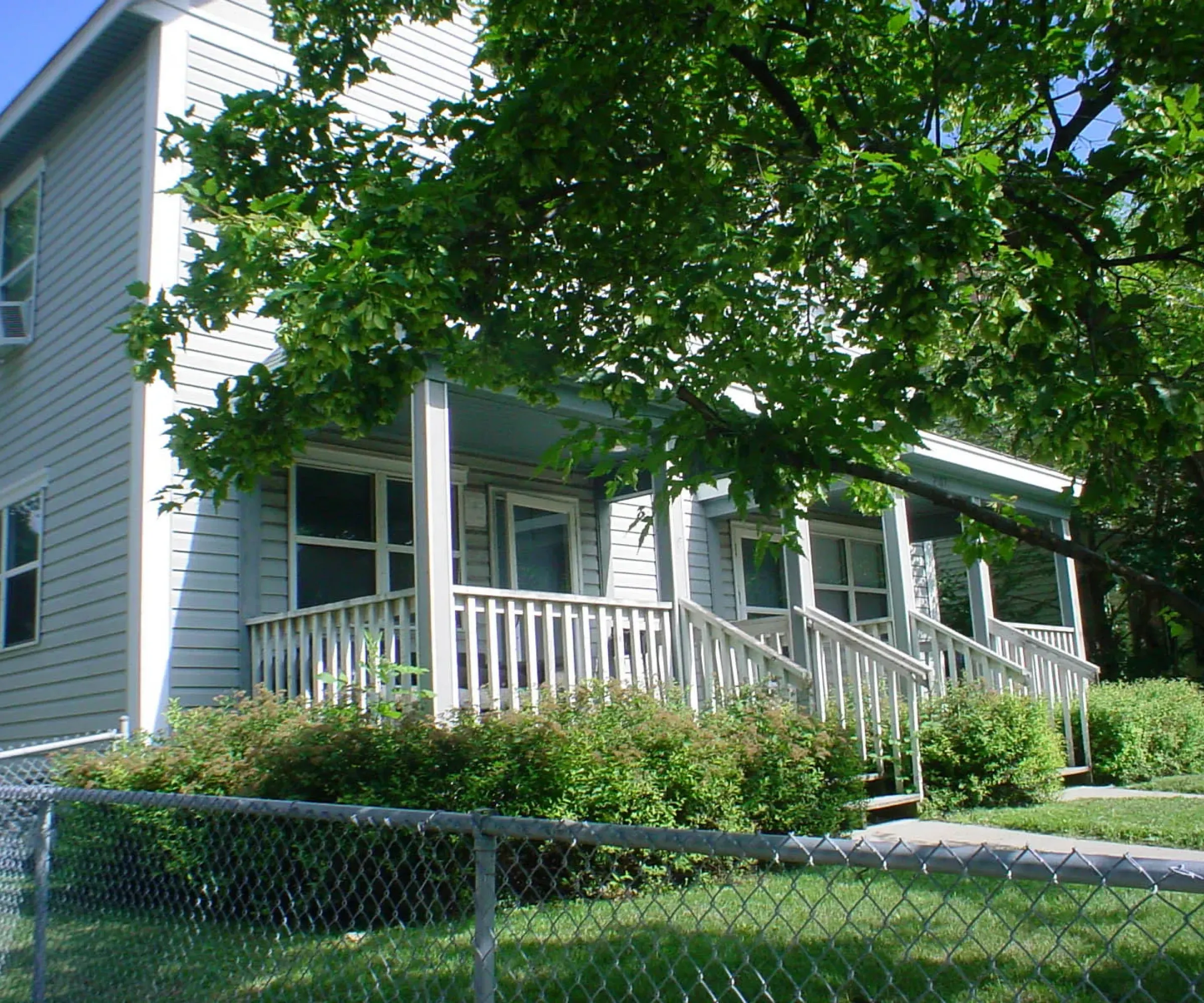Aeon Affordable Homes and Community A two-story white house with a porch, surrounded by green trees and a metal fence, offers affordable elegance on a sunny day.