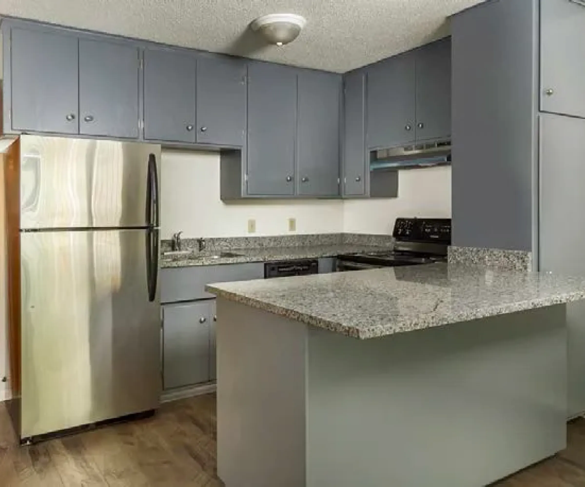 Aeon Affordable Homes Compact kitchen at Cobblestone Court Apartments with granite countertops, stainless steel fridge, gray cabinets, and a wooden floor.