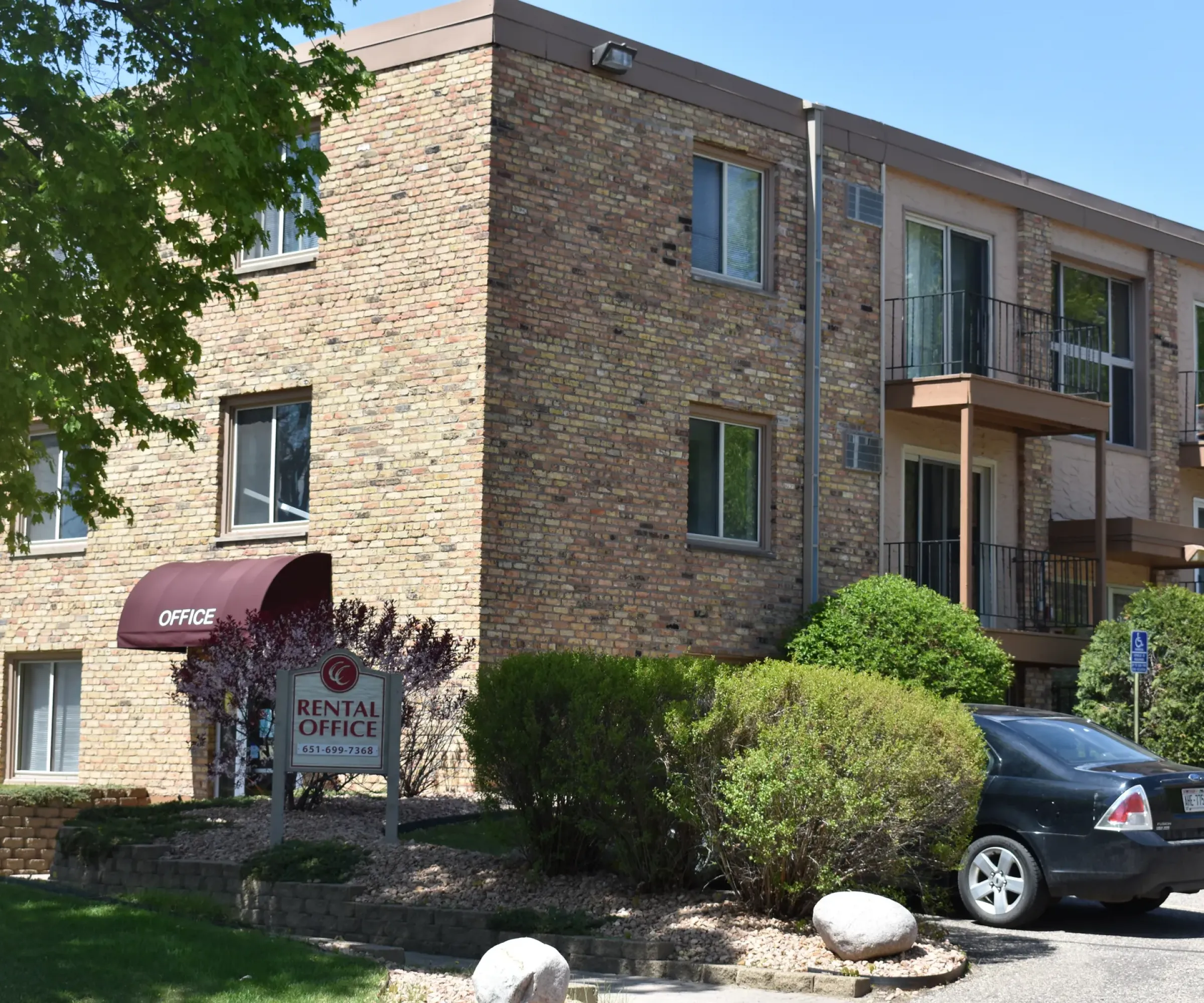 Aeon Affordable Homes Cobblestone Court Apartments features a brick facade, a rental office, lush greenery in front, and a parked car on the right.