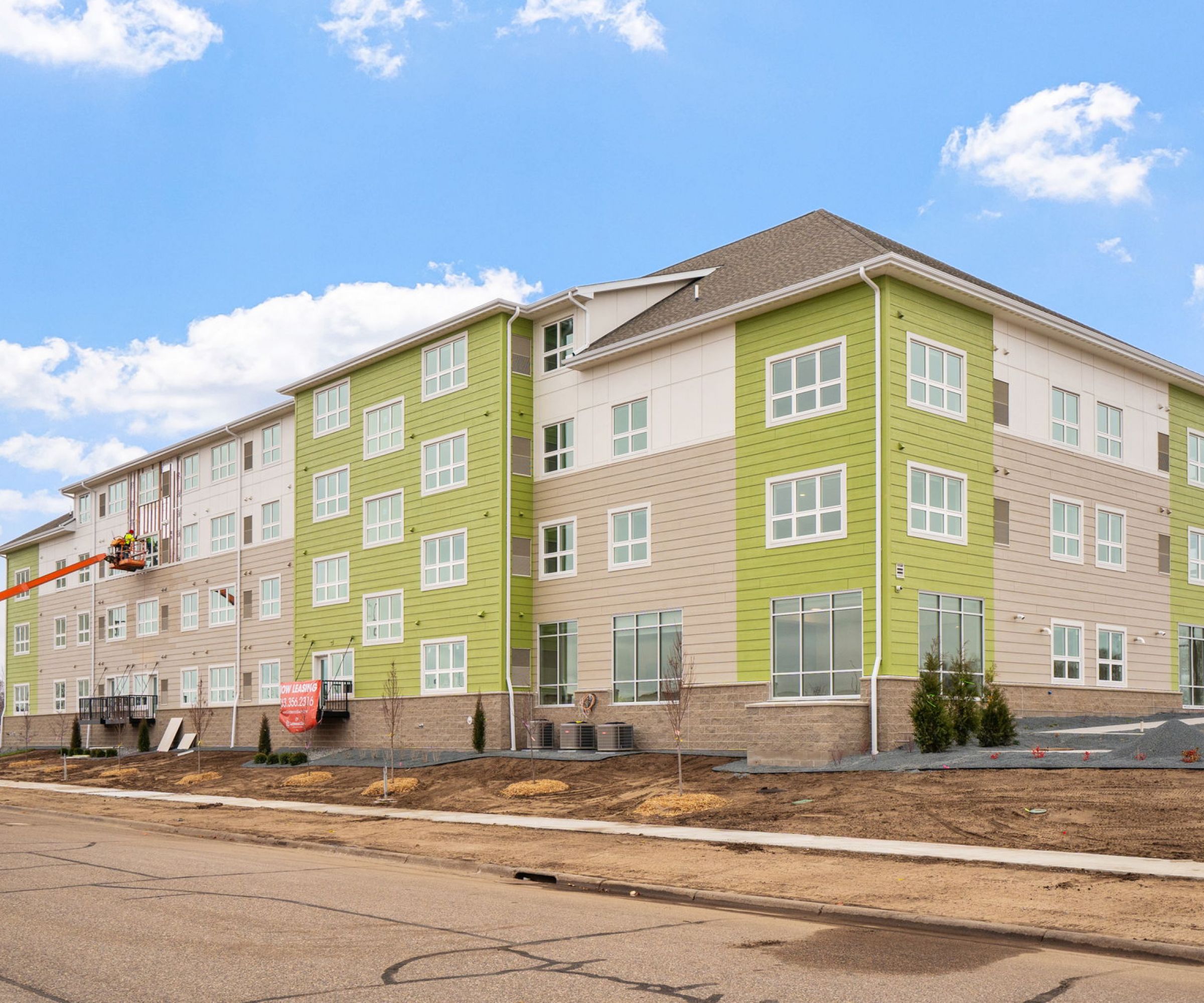 Aeon Affordable Homes and Community Four-story green and beige building under construction, promising affordable apartment homes, with workers on a lift against a clear blue sky.