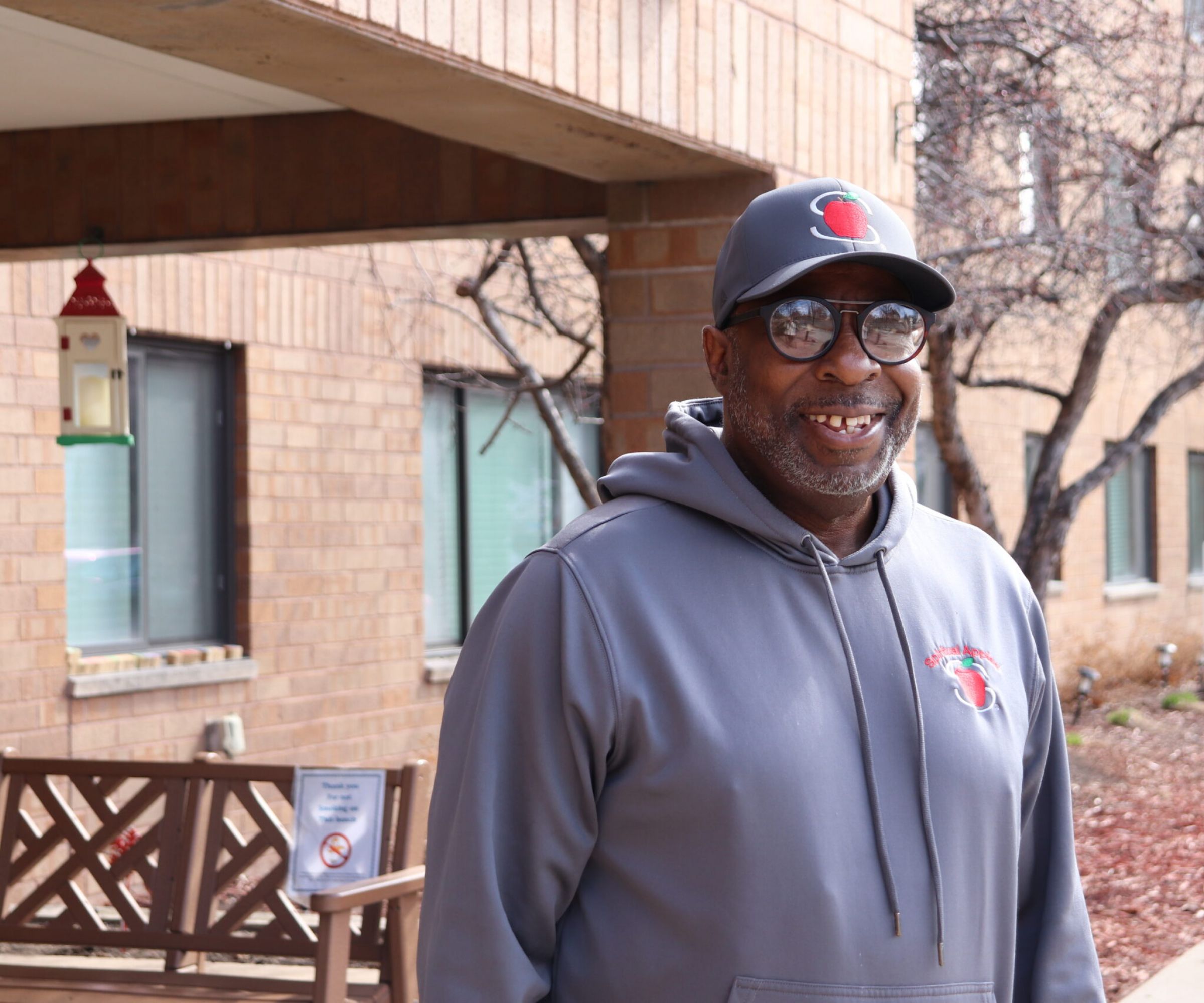 Aeon Affordable Homes Smiling man in a gray hat and hoodie stands outside a brick building, near a bench and bird feeder, sharing James's Story.