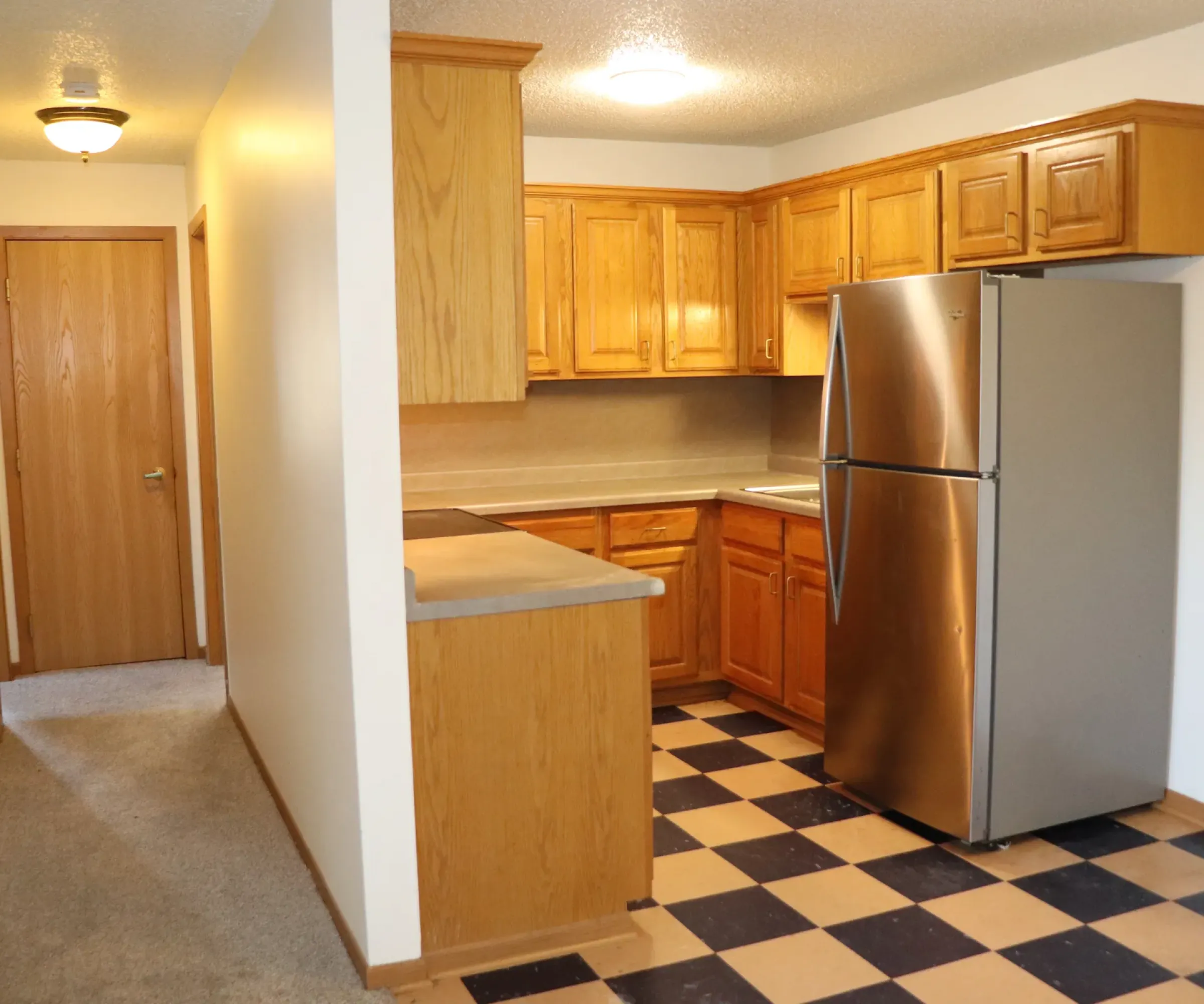 Aeon Affordable Homes and Community Kitchen with wooden cabinets, stainless steel refrigerator, and checkered floor in affordable apartment homes. Hallway to the left with carpet.