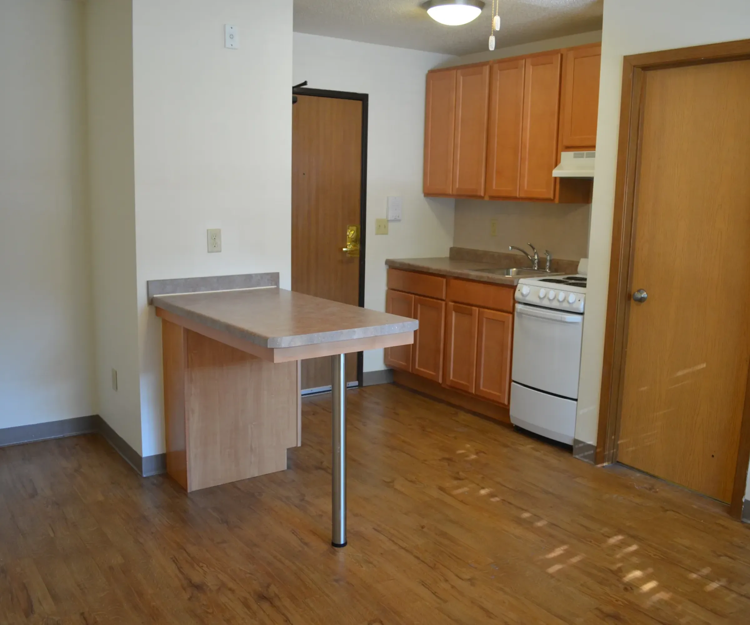 Aeon Affordable Homes and Community Affordable apartment homes featuring a compact kitchen with wooden cabinets, a small stove, and an elevated counter. Wood flooring throughout.
