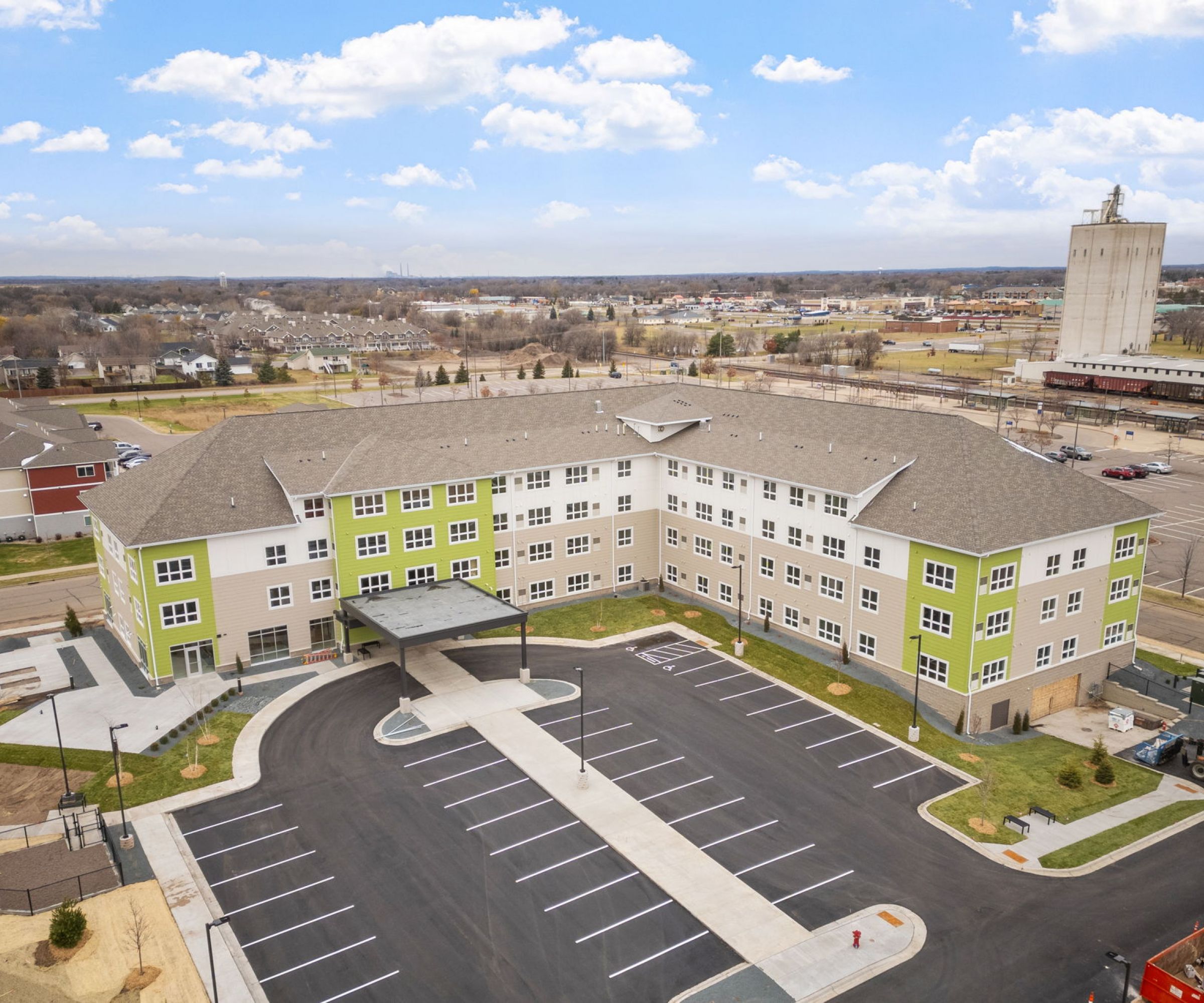 Aeon Affordable Homes and Community Aerial view of a large, L-shaped building with green accents, offering affordable apartment homes, surrounded by parking and open landscape.