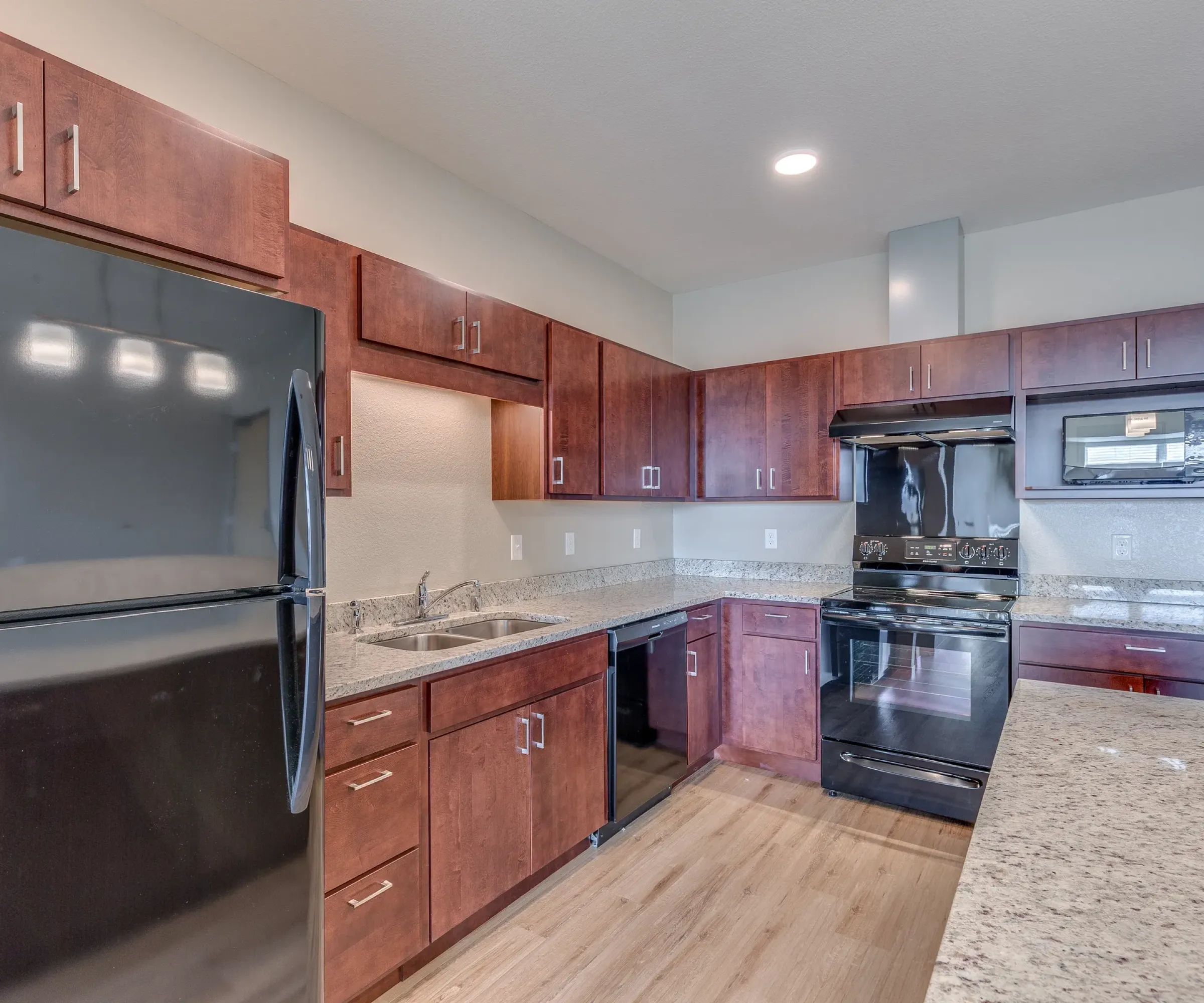 Aeon Affordable Homes and Community Modern kitchen in affordable apartment homes with dark wooden cabinets, granite countertops, black appliances, and light wood flooring.