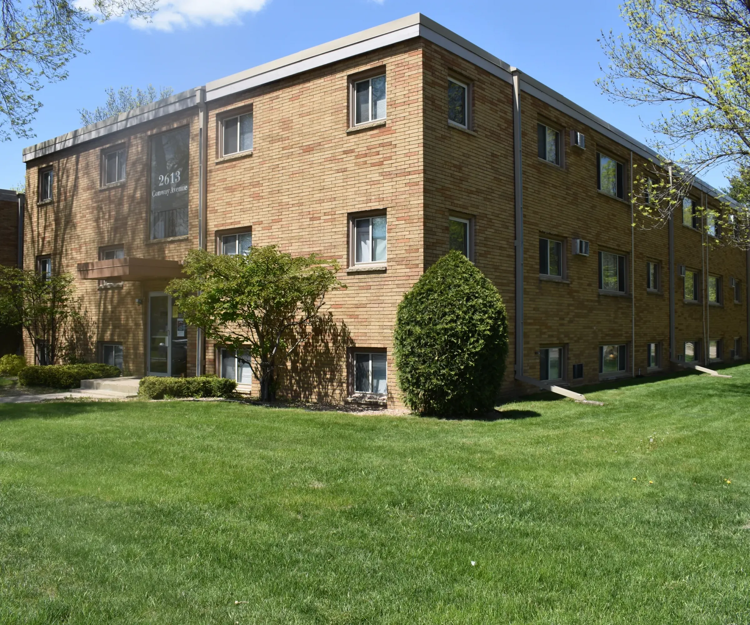 Aeon Affordable Homes Cobblestone Court Apartments: A brick building with rectangular windows, nestled amid grass and trees under a clear blue sky.
