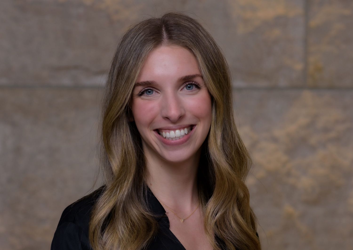 Aeon Affordable Homes Rachel Azorsky, with long, wavy blonde hair, smiles in a black top against a stone wall background.