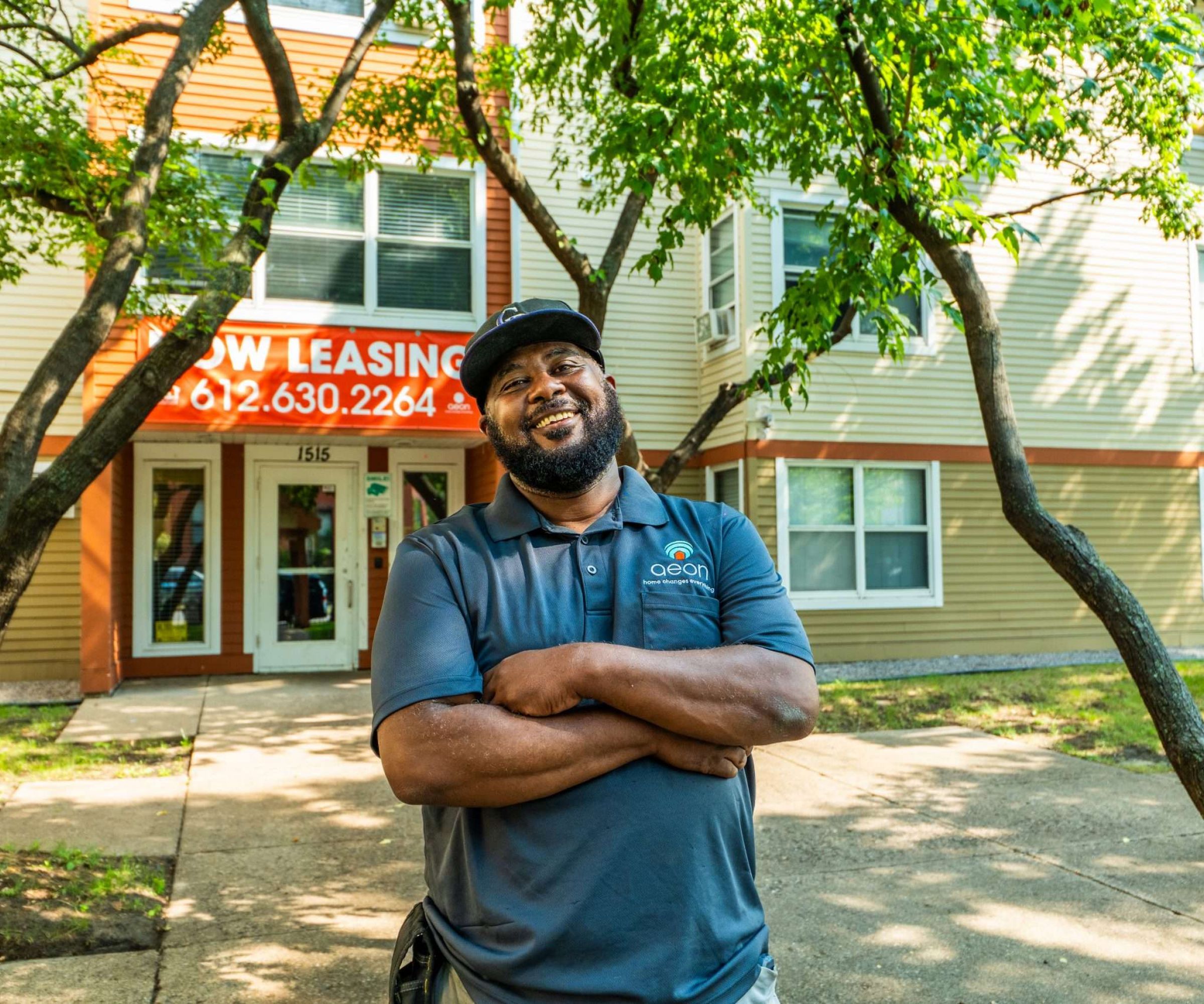 Aeon Affordable Homes and Community Man smiling with arms crossed in front of an apartment building with "Now Leasing" sign.