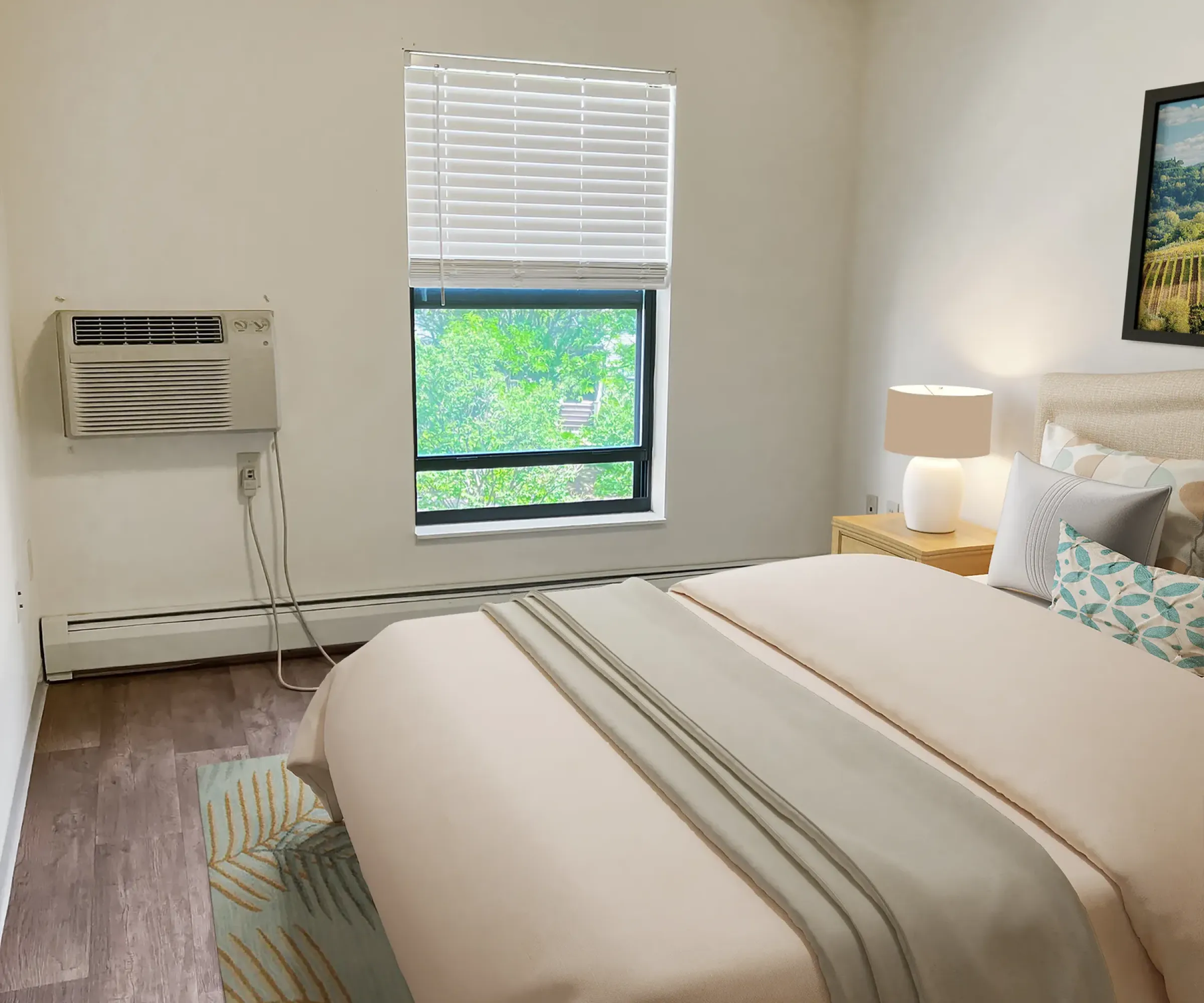 Aeon Affordable Homes and Community Cozy bedroom in affordable apartment homes with a bed, lamp, wall art, and a window view of greenery. Wall-mounted air conditioner included.