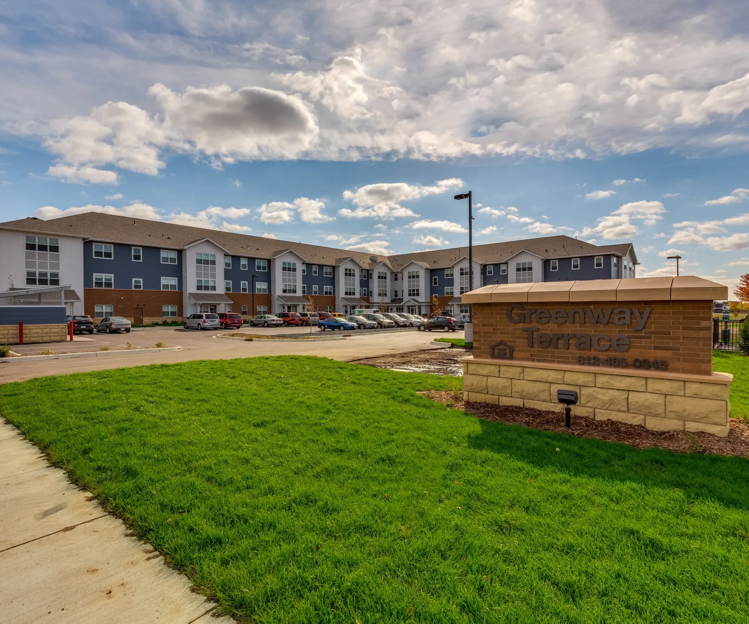 Aeon Affordable Homes and Community Affordable apartment homes at "Greenway Terrace" are surrounded by lush grass, under a partly cloudy sky.