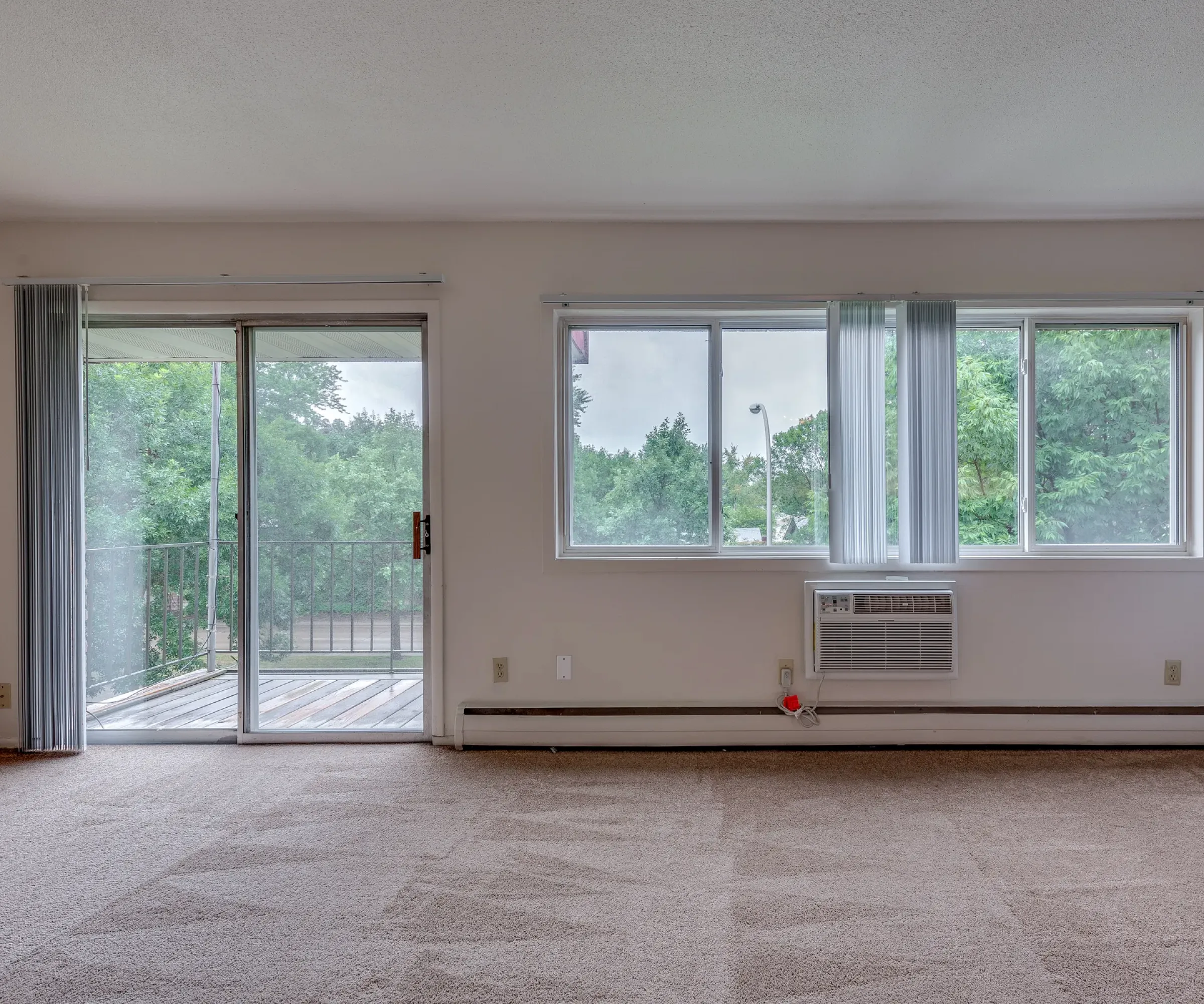 Aeon Affordable Homes and Community Affordable apartment home with beige carpet, a large window, air conditioner, and sliding door leading to a balcony with lush greenery.