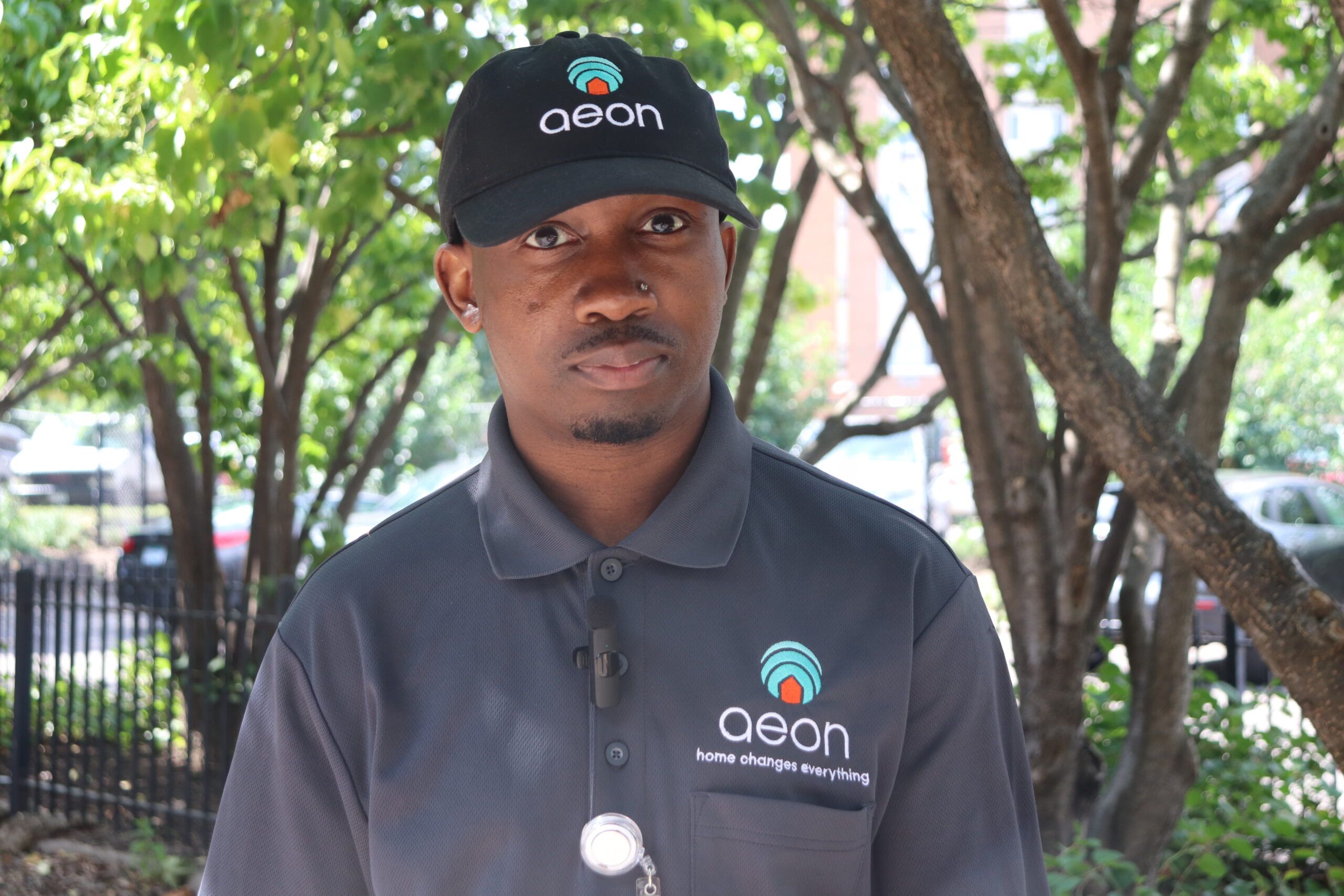 Aeon Affordable Homes A man in an "Aeon" uniform and hat stands outside in front of trees and a black fence.