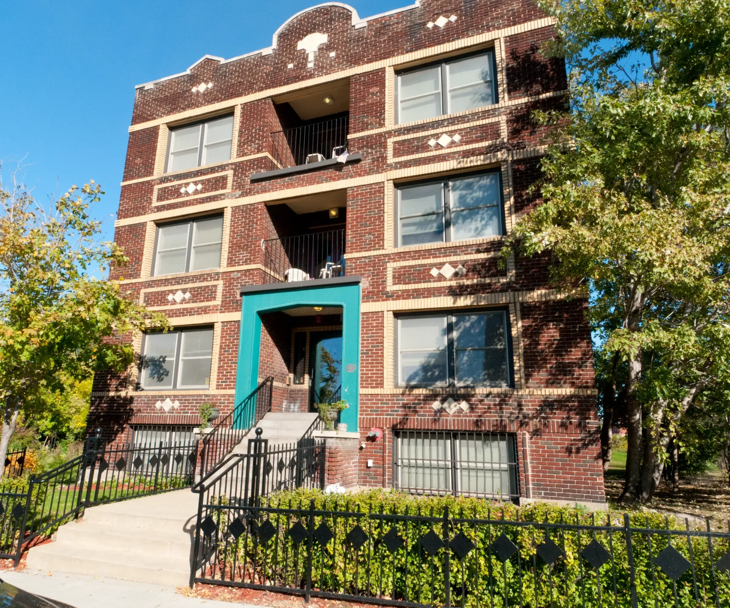 Aeon Affordable Homes and Community A four-story red brick building, offering affordable apartment homes, is adorned with decorative patterns and surrounded by trees under a clear blue sky.