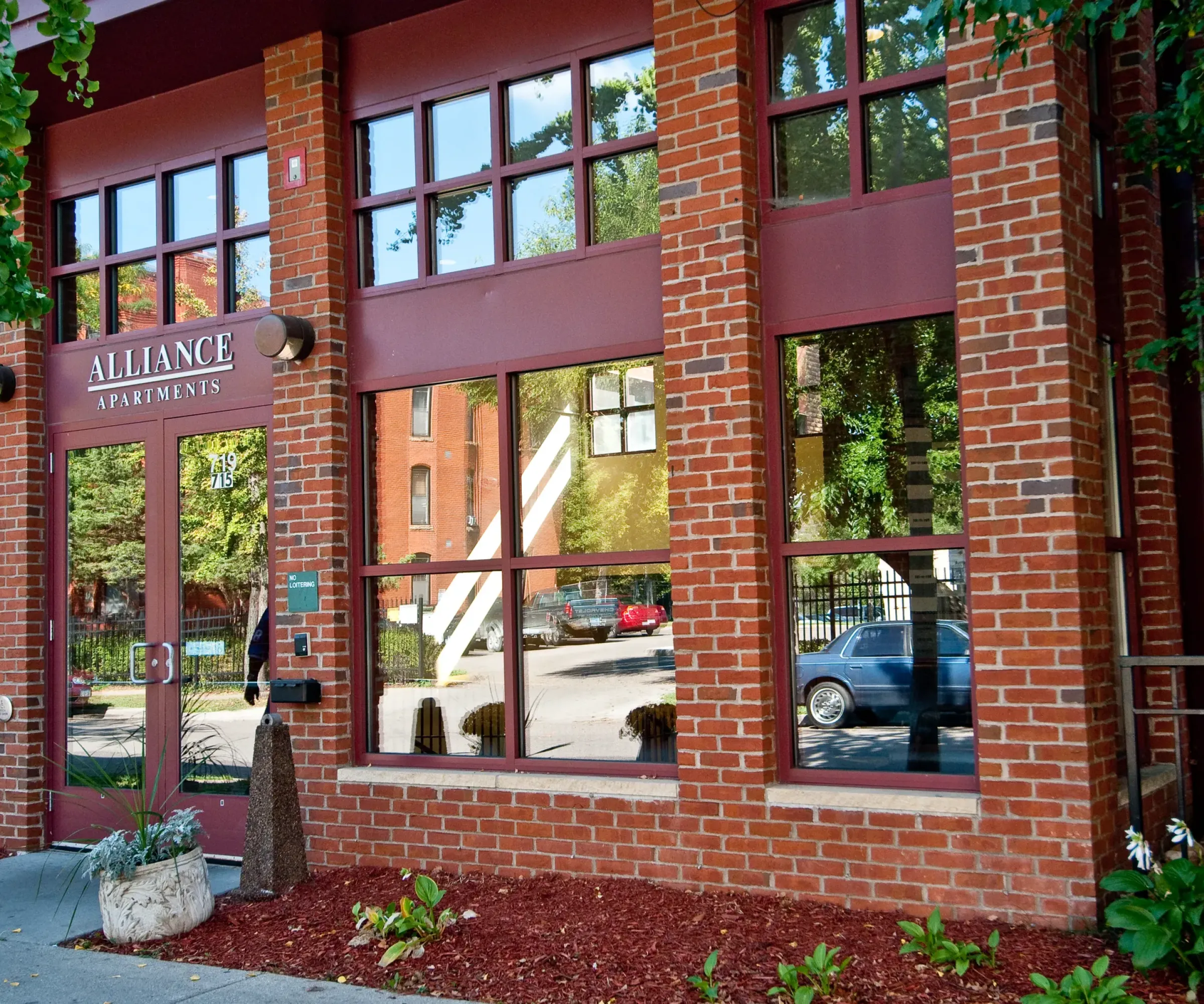 Aeon Affordable Homes and Community Brick building with large windows, entrance labeled "Alliance Apartments," featuring a small garden area in front. Discover affordable apartment homes today.