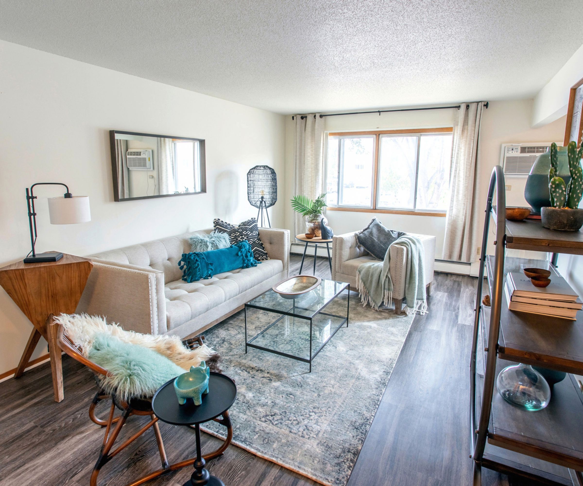 Aeon Affordable Homes Cozy living room with a beige sofa, accent chairs, shelves, a small table, and a large window with light curtains.