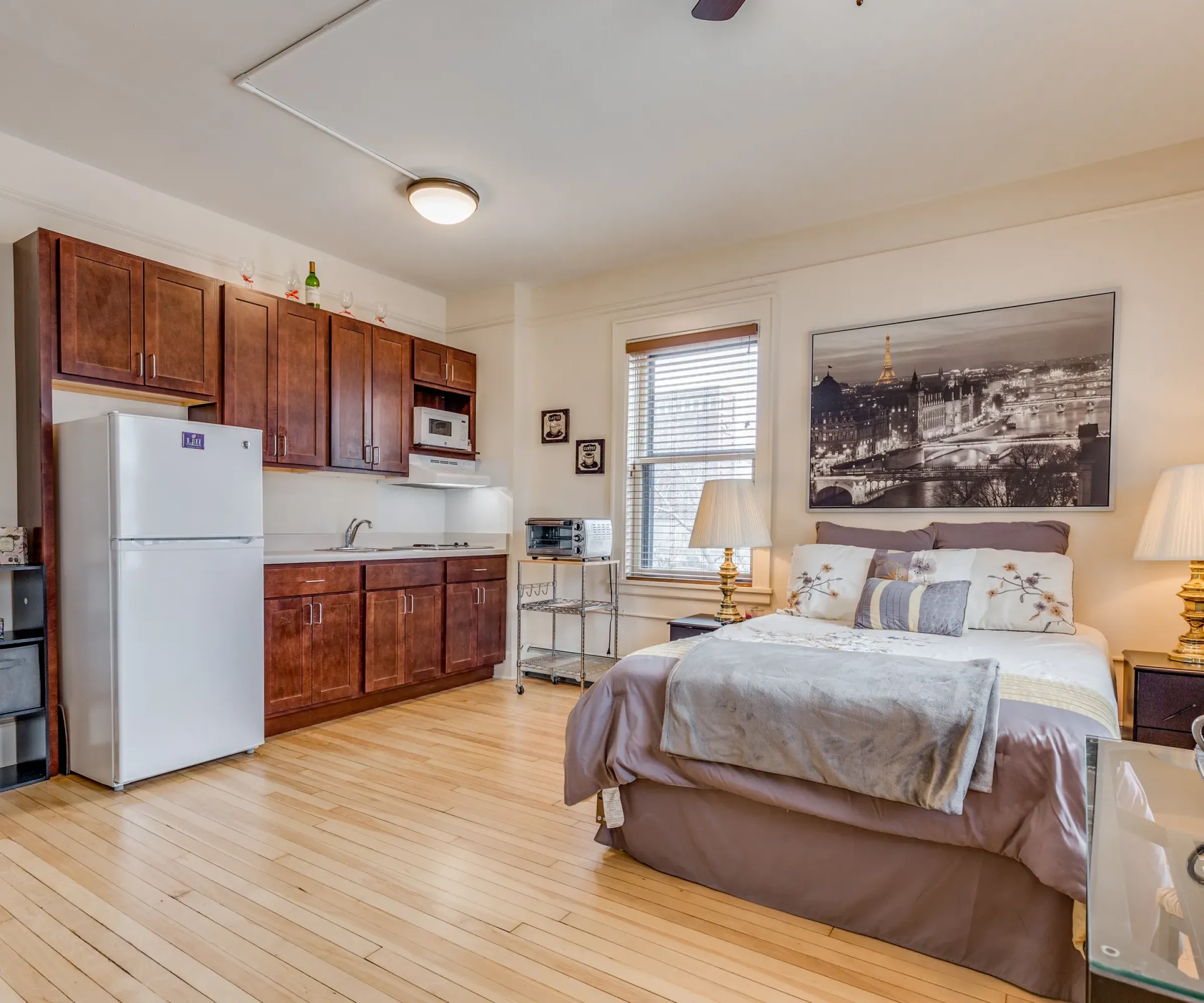 Aeon Affordable Homes and Community Affordable studio apartment with kitchenette, bed, and chic decor. Wooden floors, lamps, and a cityscape photo on the wall.
