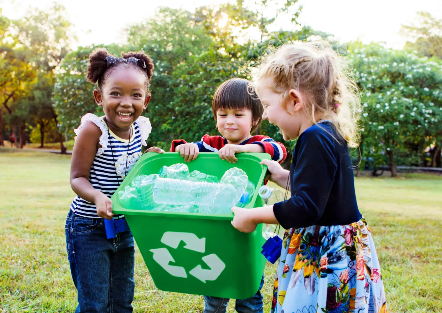 Aeon Affordable Homes with three children smiling and holding a green recycling bin filled with plastic bottles in a grassy park.