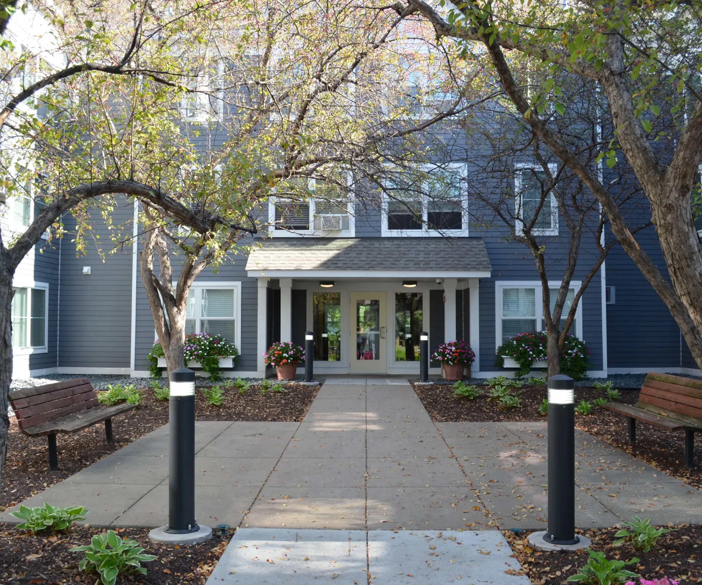Aeon Affordable Homes and Community A path leads to the entrance of affordable apartment homes, flanked by trees and benches, with potted plants near the door.