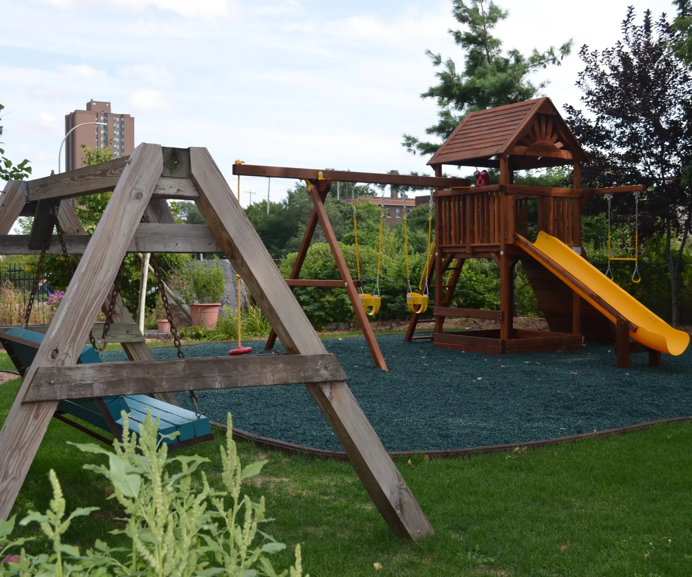 Aeon Affordable Homes and Community Children's playground with swings, a wooden playset, and a yellow slide, nestled near affordable apartment homes in lush greenery.