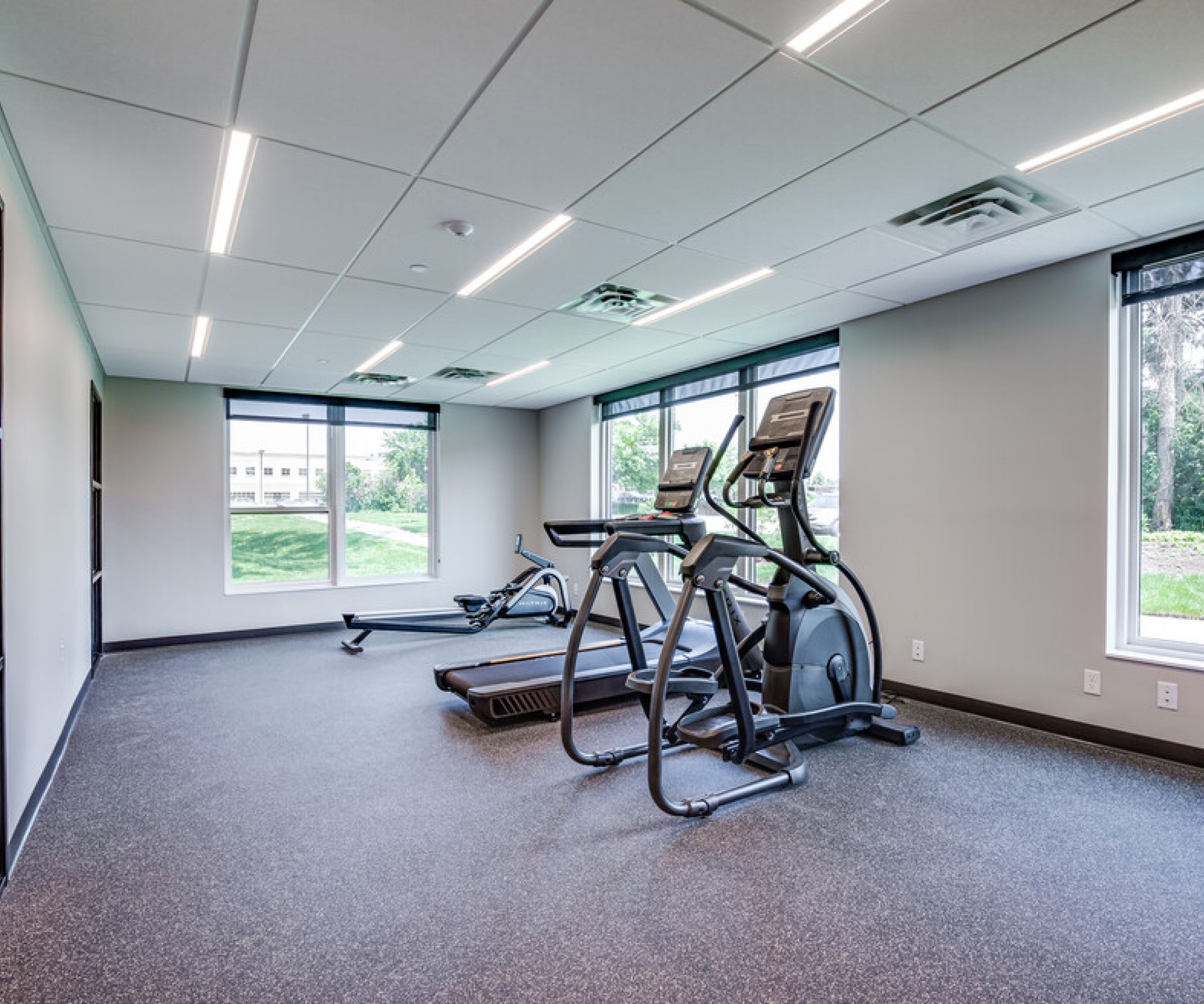 Aeon Affordable Homes and Community Affordable apartment homes featuring a small gym with two treadmills, an exercise bike, and large windows overlooking greenery. Modern, well-lit space.