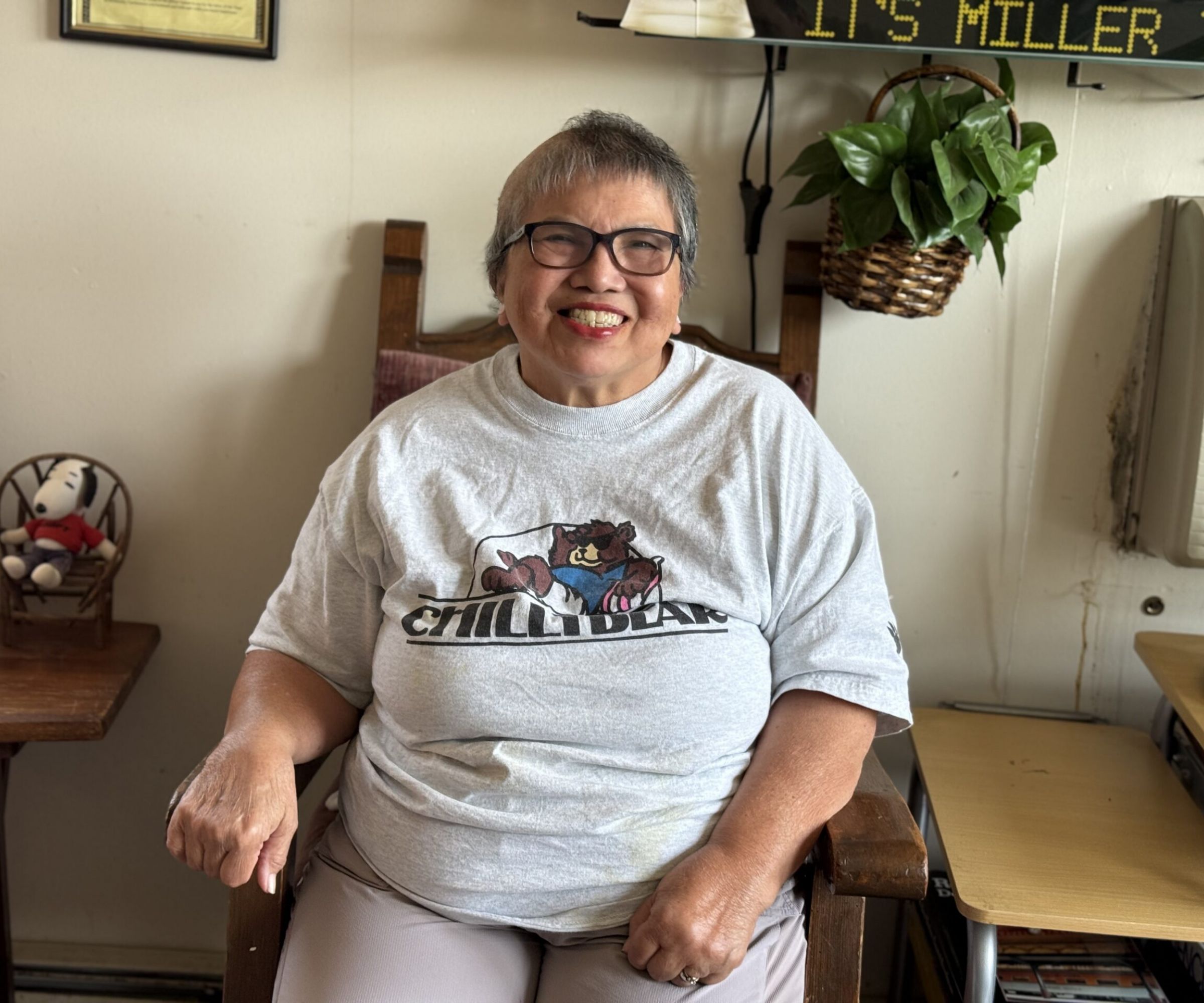 Aeon Affordable Homes Smiling woman with short gray hair sits indoors wearing glasses and a gray “CHILLI BEAR” T-shirt, sharing Amelia's Story.