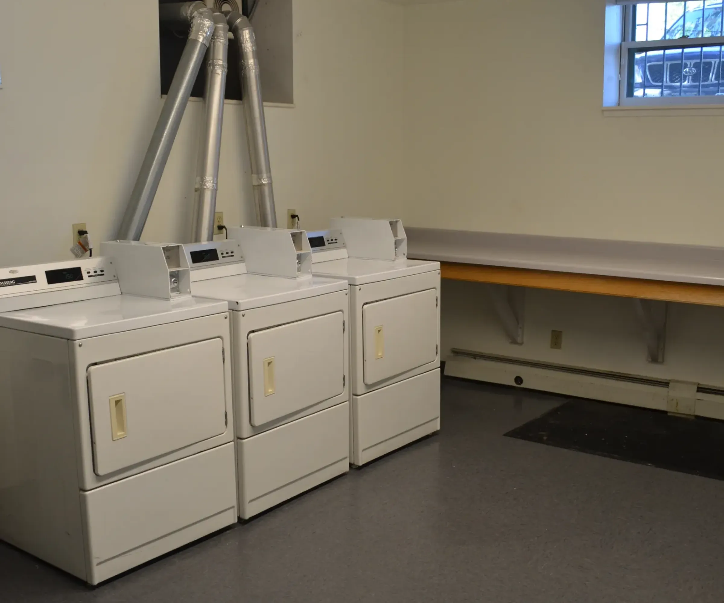 Aeon Affordable Homes and Community Affordable apartment homes feature a laundry room with three dryers, a folding table, and a small window. The floor is gray, and the walls are light-colored.