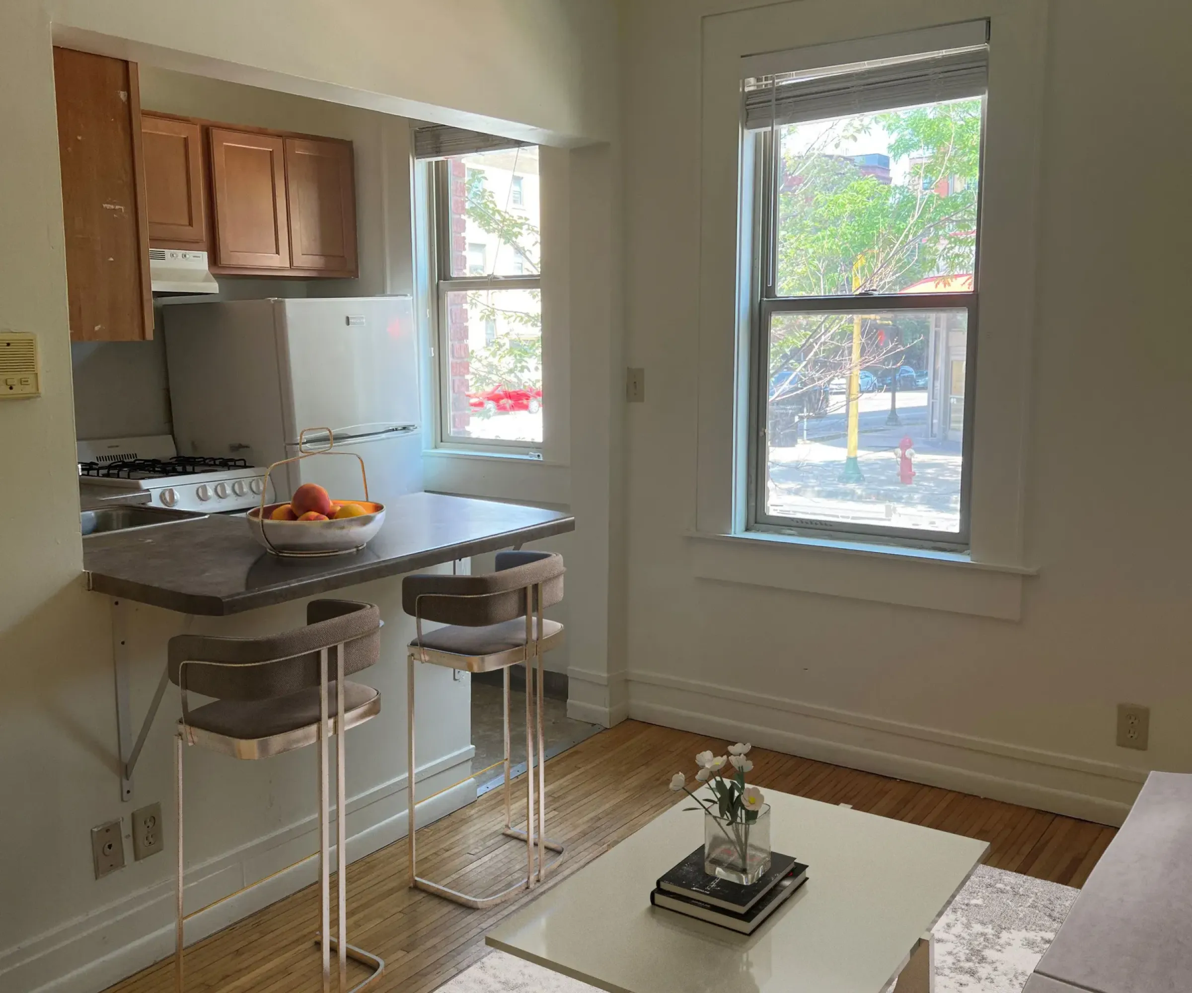 Aeon Affordable Homes and Community Cozy living room in affordable apartment homes with a white table, gray couch, and two bar stools by the kitchen counter near bright windows.
