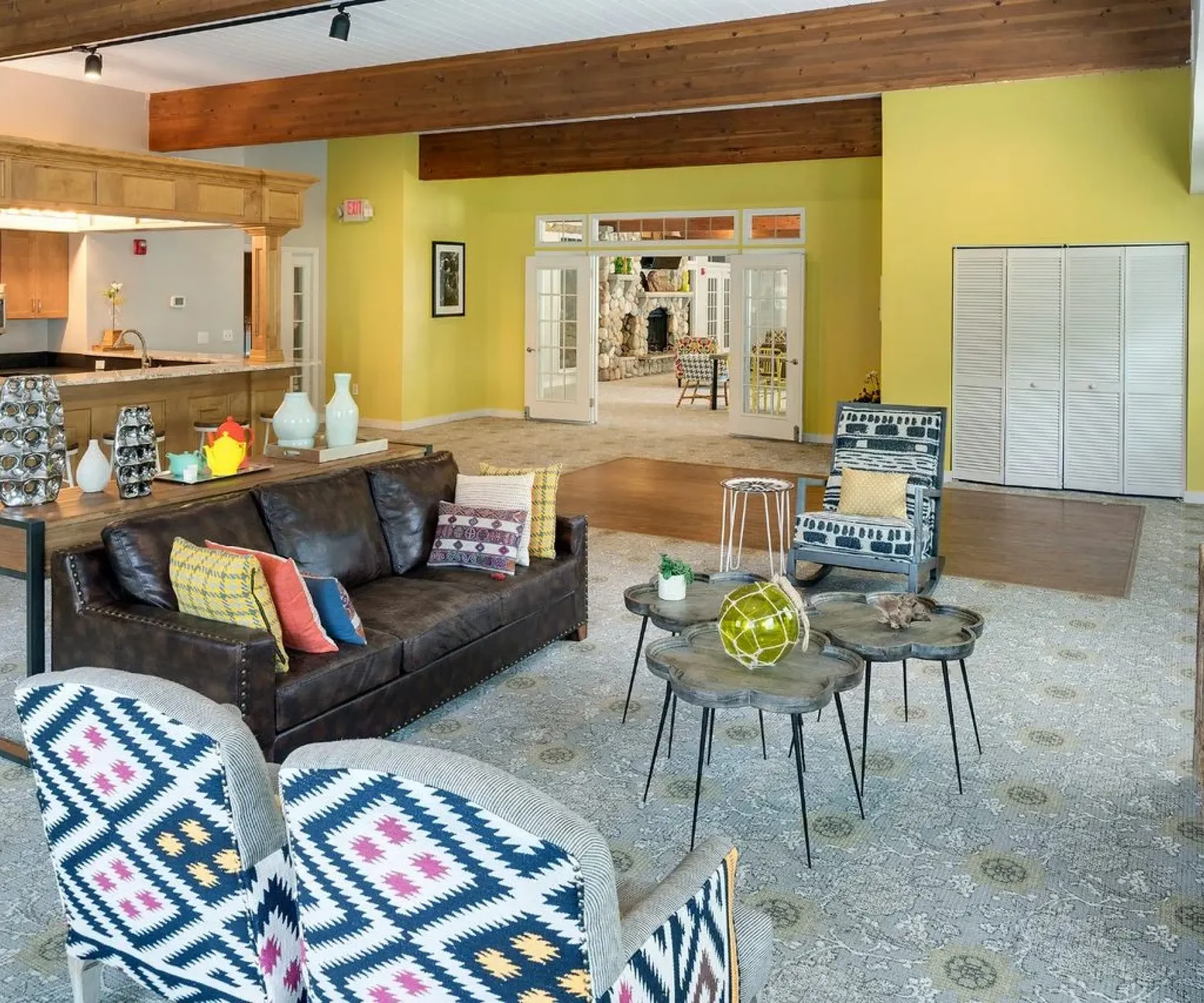 Aeon Affordable Homes Modern living area in Blooming Meadows with patterned chairs, brown sofa, wooden tables, and bright yellow walls. Open kitchen in background.