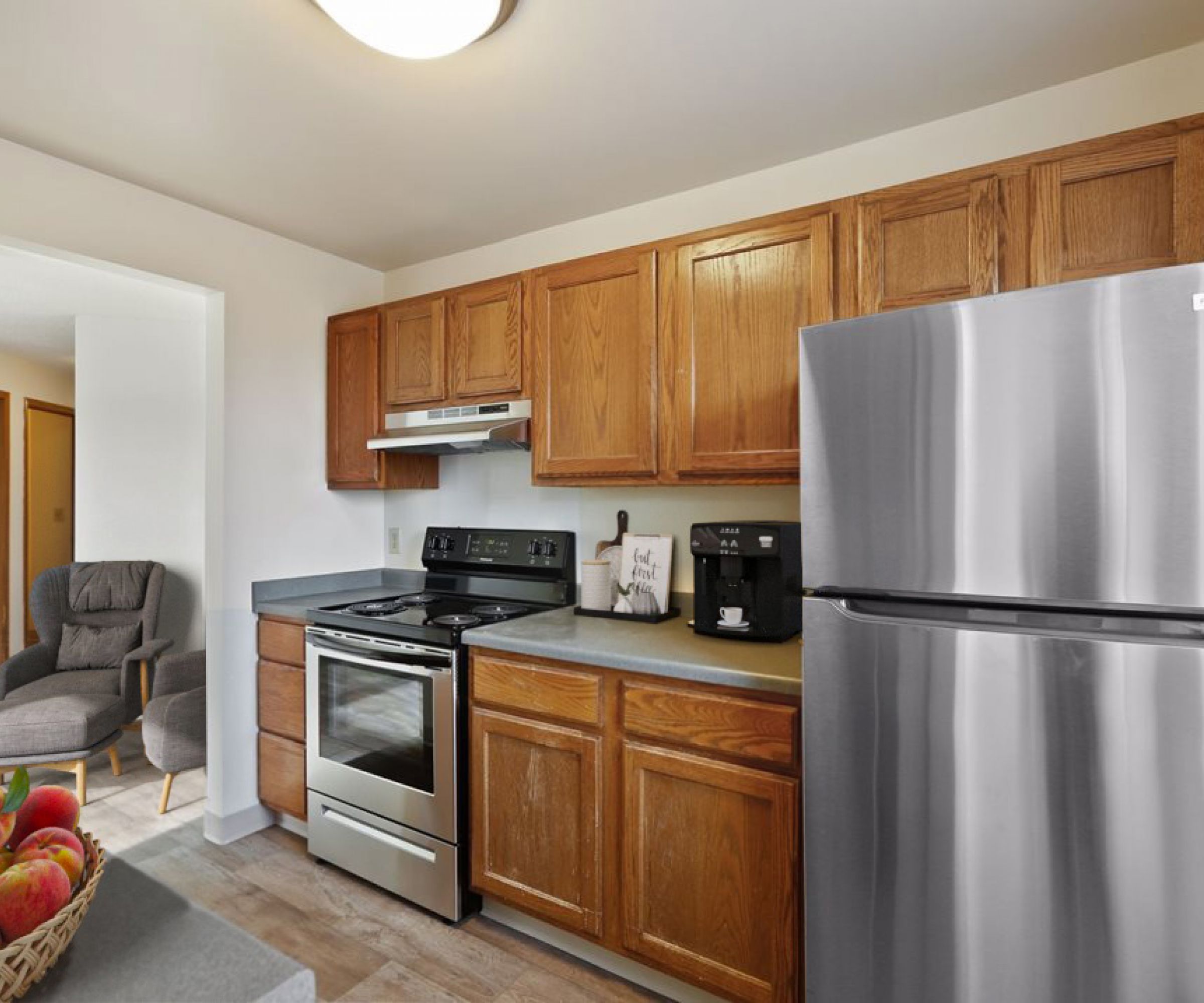 Aeon Affordable Homes and Community Modern kitchen in affordable apartment homes with wooden cabinets, stainless steel appliances, a bowl of apples, and a chair nearby.