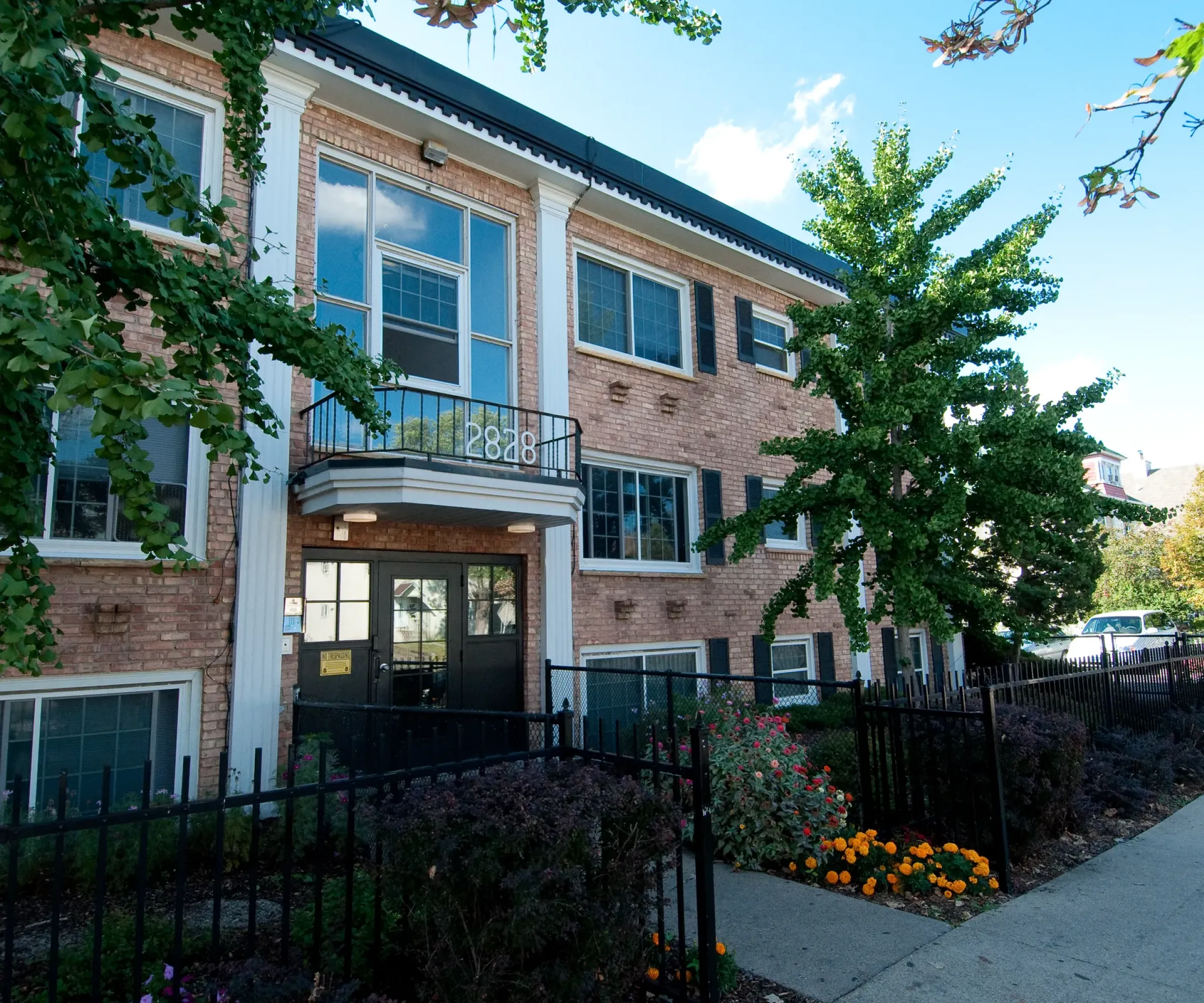 Aeon Affordable Homes and Community Affordable apartment homes in a red brick building with black doors and windows, nestled amidst greenery and flowers under a blue sky.