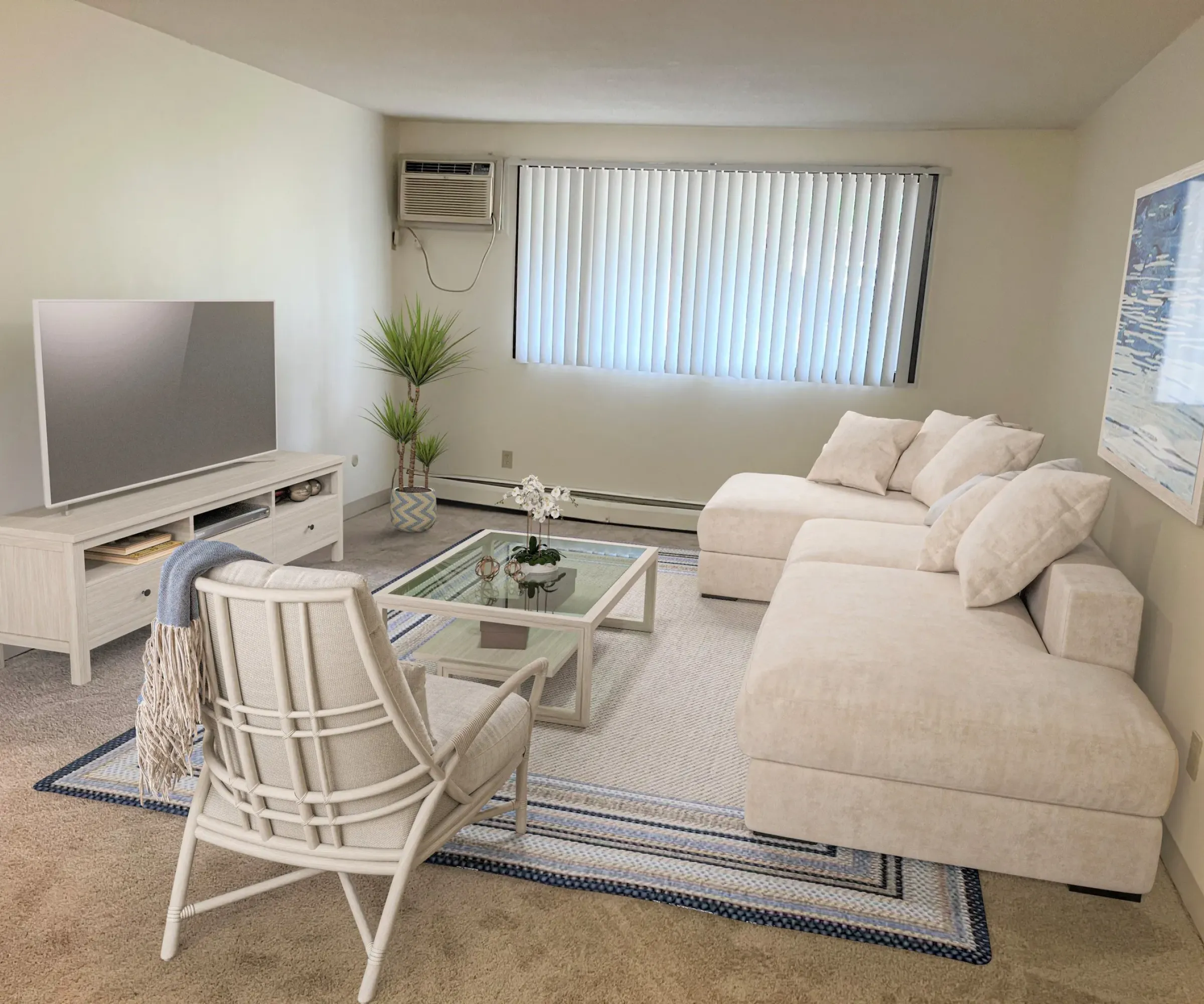 Aeon Affordable Homes and Community Modern living room in affordable apartment homes, featuring a white sectional sofa, glass coffee table, TV, and large window with blinds.