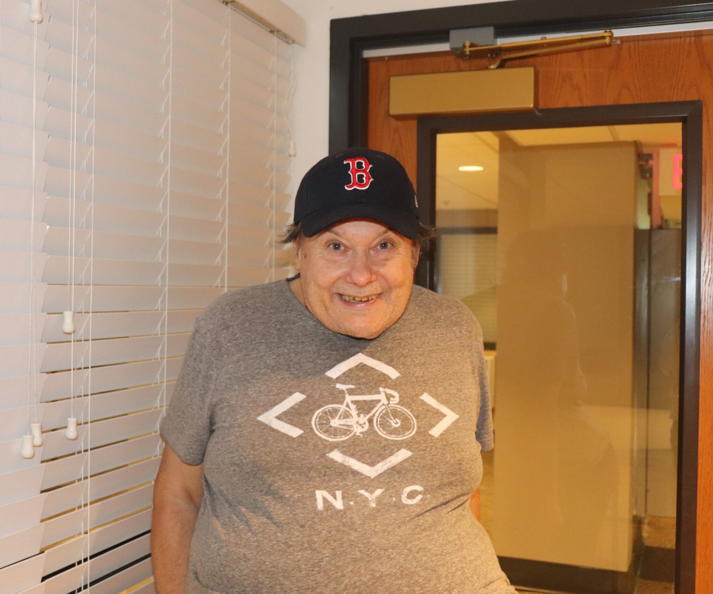 Aeon Affordable Homes Smiling elderly man in a Boston Red Sox hat and NYC bike t-shirt stands indoors by a door and window blinds.