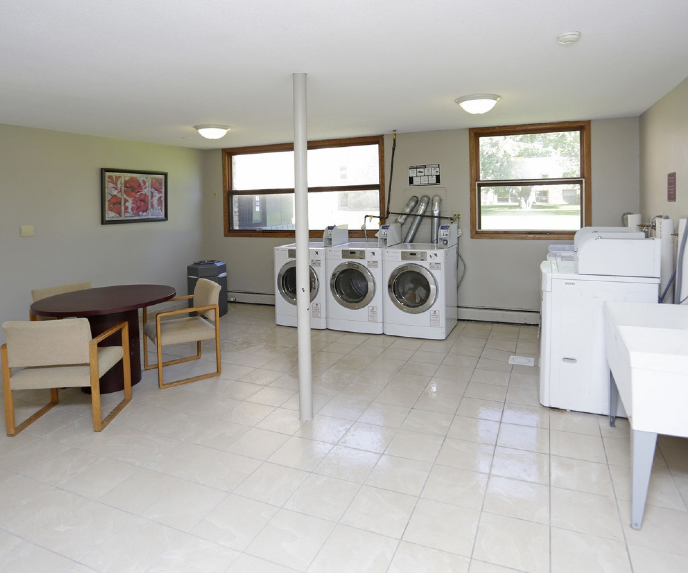 Aeon Affordable Homes and Community Affordable apartment homes feature a laundry room with washers, dryers, a sink, and a small dining table. Large windows let in natural light.