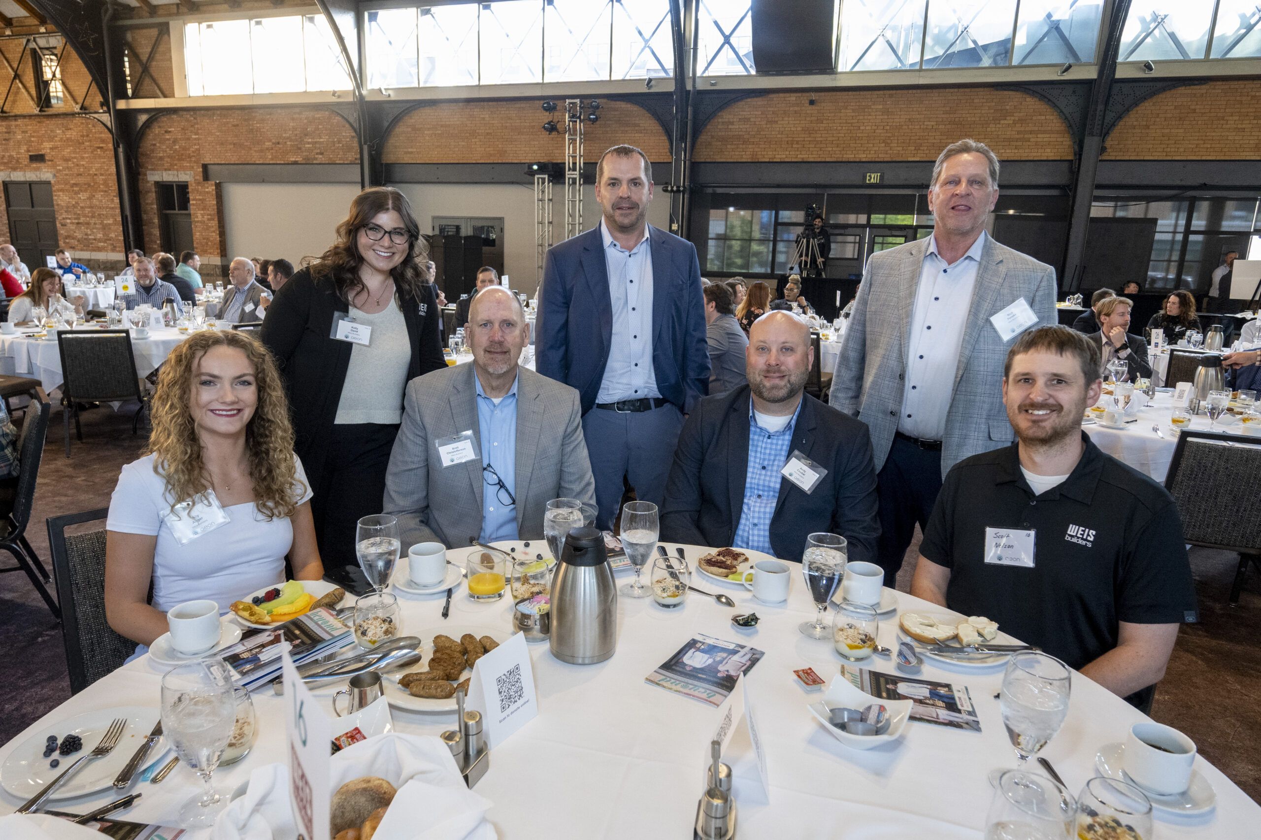 Aeon Affordable Homes Seven people pose around a table at a formal breakfast event in a large, bright venue.