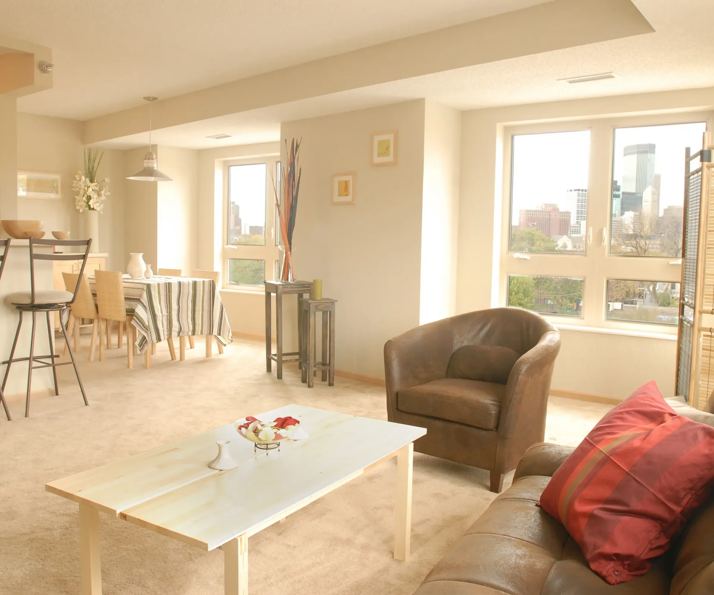 Aeon Affordable Homes and Community Bright living room in affordable apartment homes with a sofa, armchair, coffee table, and dining area overlooking a cityscape.