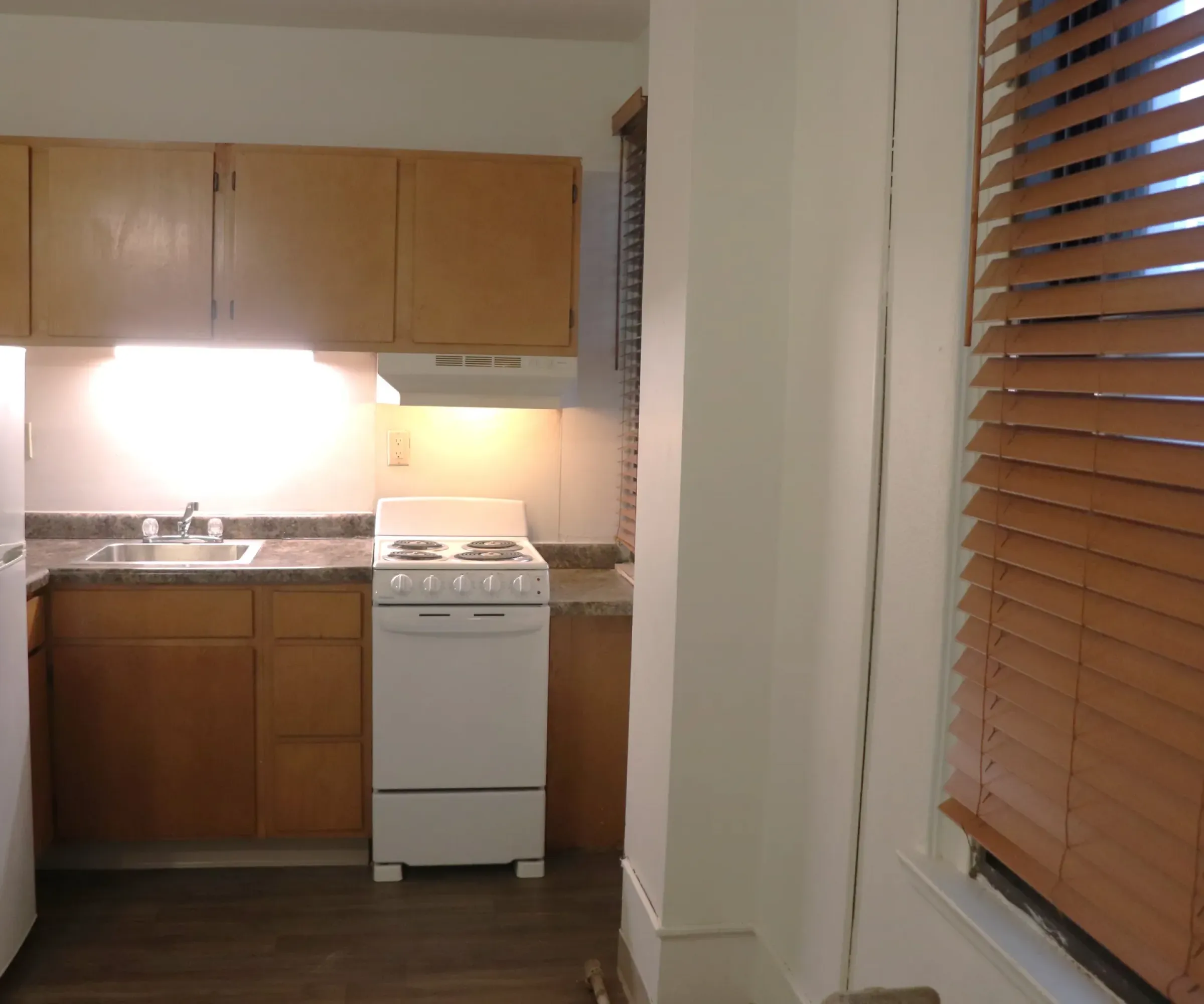 Aeon Affordable Homes and Community Affordable apartment homes with a small kitchen, featuring a fridge, stove, wooden cabinets, and blinds.