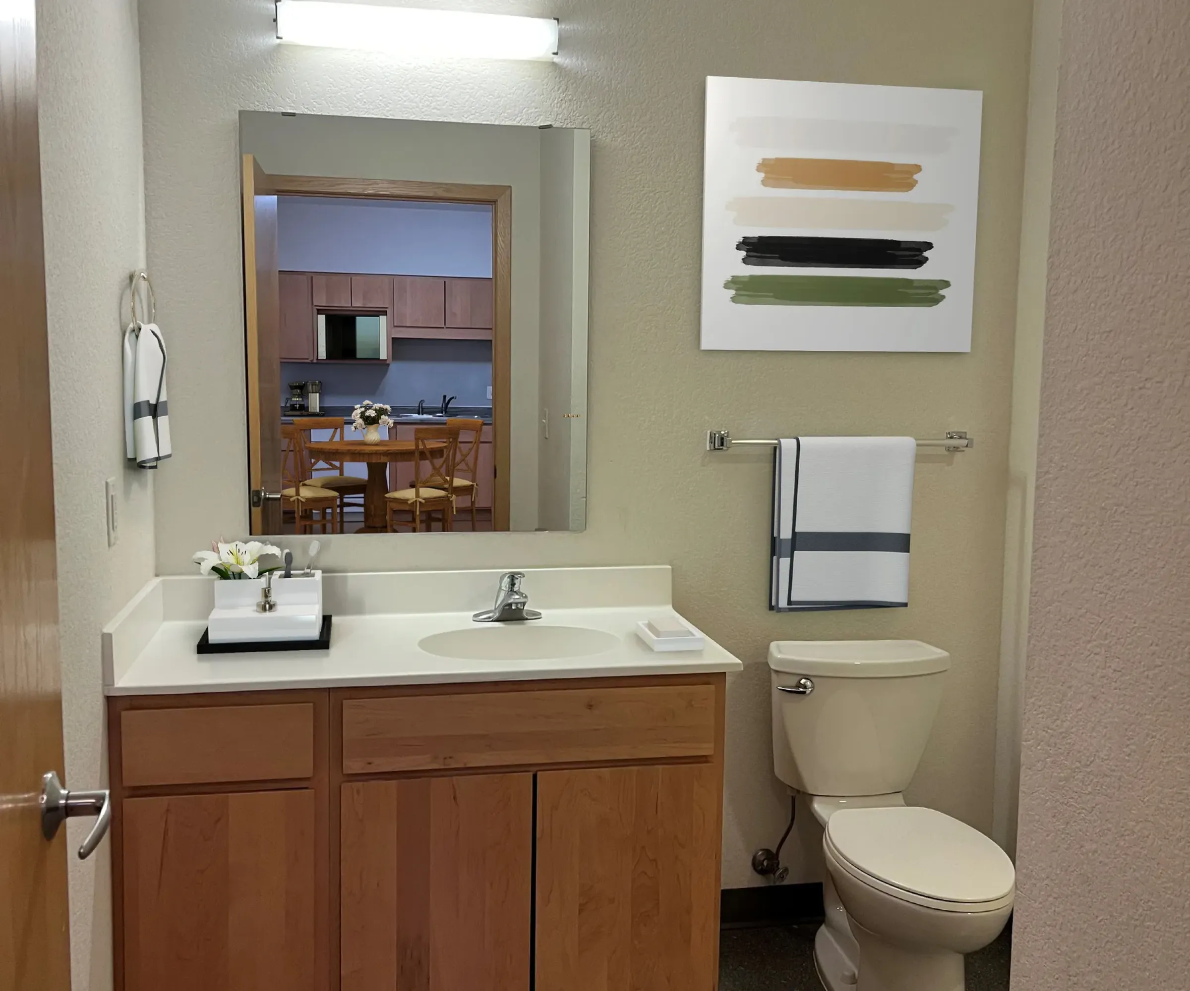 Aeon Affordable Homes and Community Modern bathroom in affordable apartment homes with wooden vanity, mirror, overhead light, toilet, abstract wall art, and a view into the kitchen.