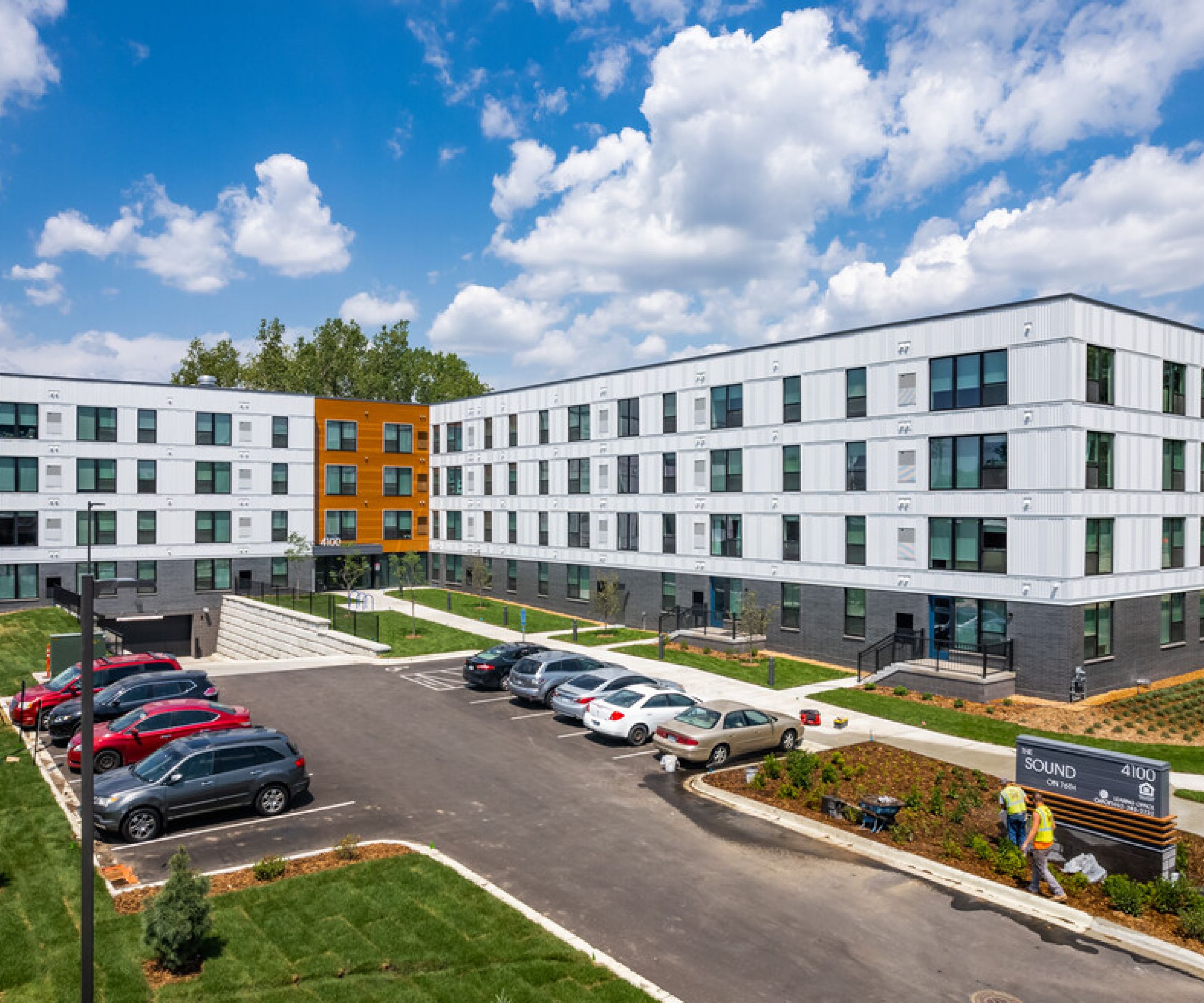 Aeon Affordable Homes and Community A modern complex of affordable apartment homes with parked cars, landscaped areas, and a clear blue sky in the background.