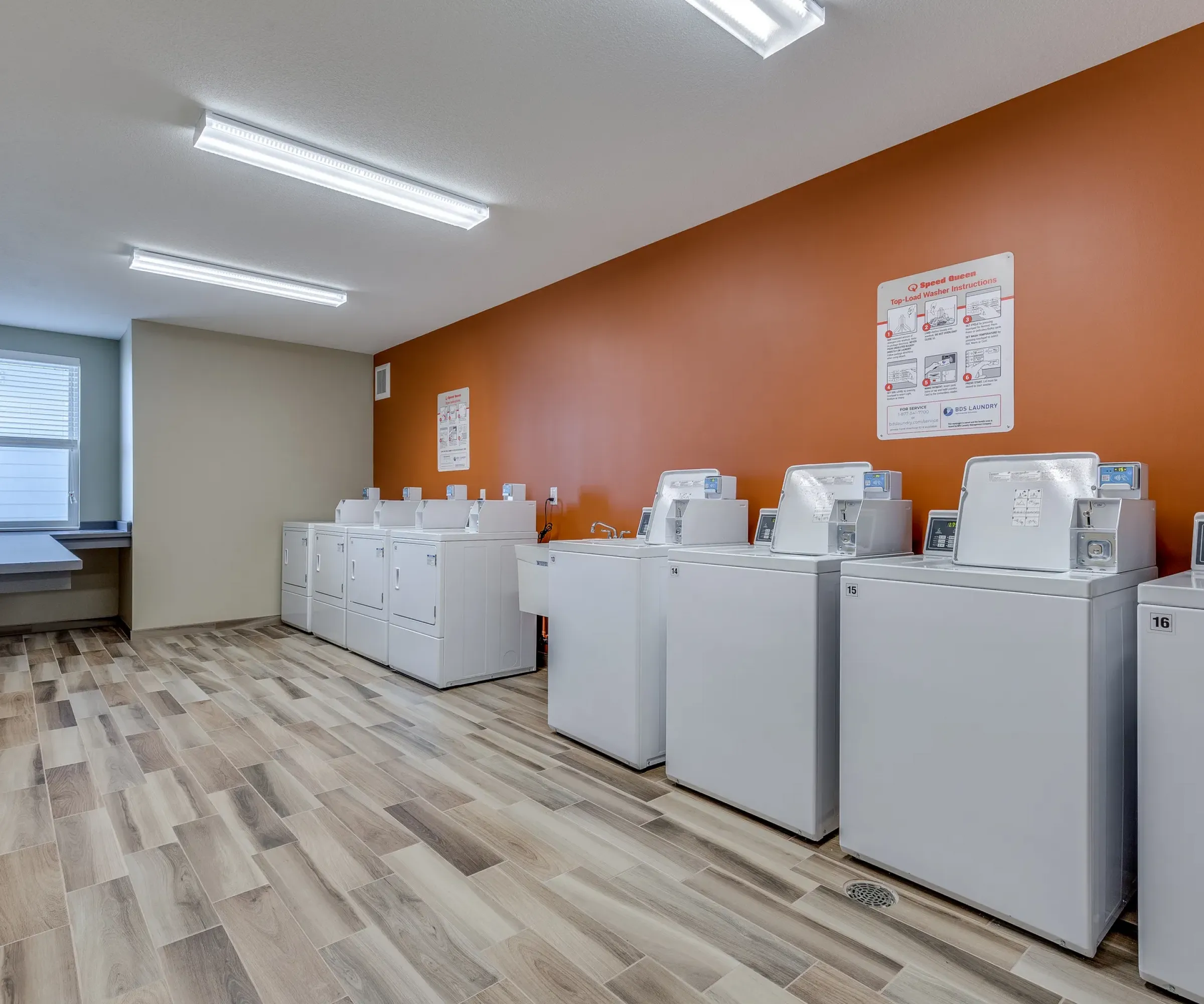 Aeon Affordable Homes and Community Laundry room in our affordable apartment homes, featuring multiple washing machines, dryers against an orange wall, and a folding table by the window.