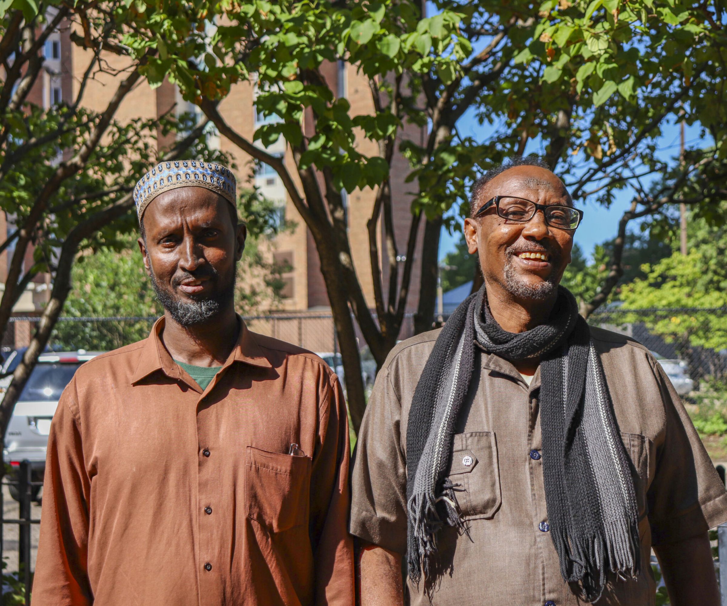 Aeon Affordable Homes Ahmed & Hassan stand outside, smiling, as their story unfolds amid trees and a sunlit building in the background.