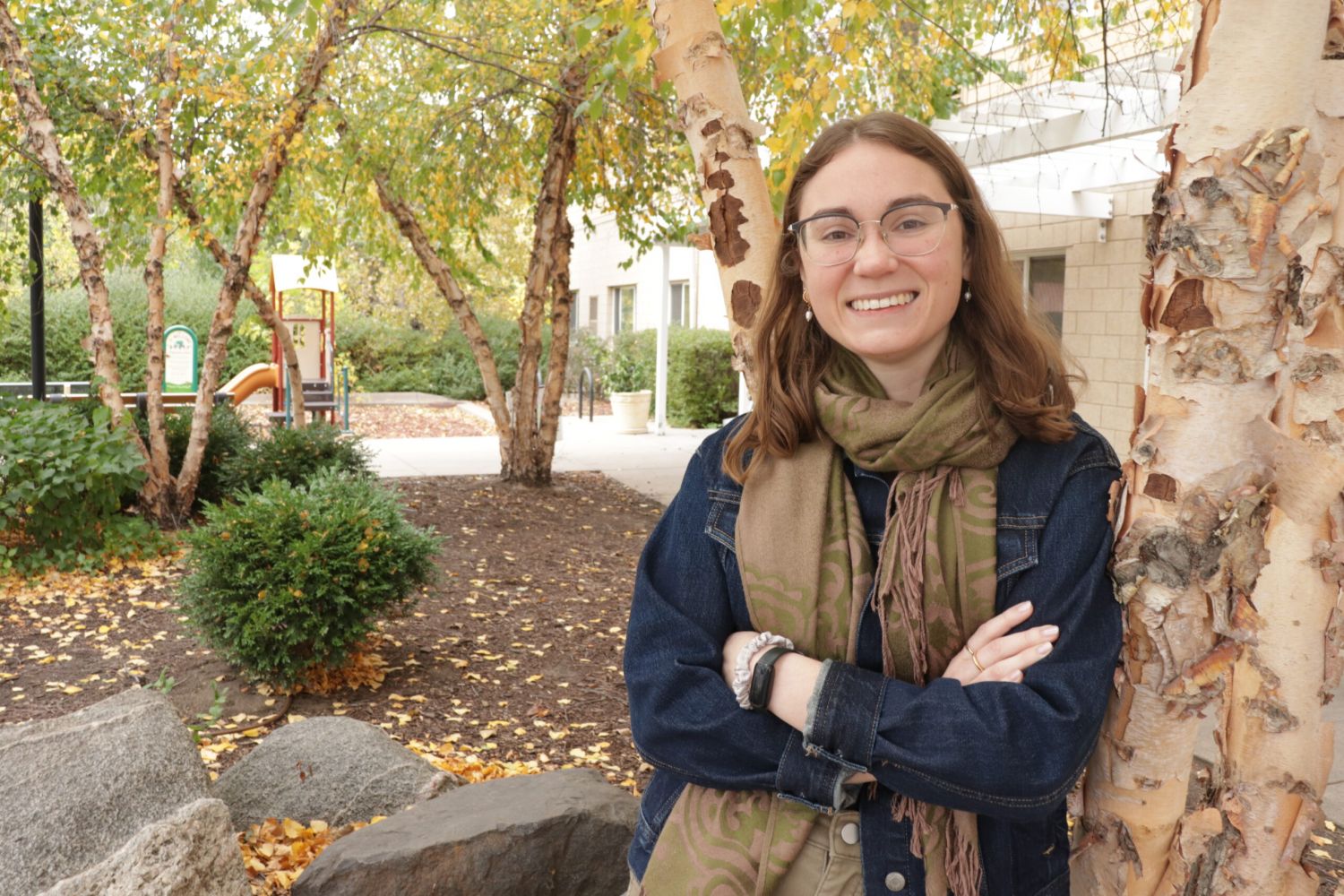Aeon Affordable Homes A smiling woman in glasses and a scarf stands by a birch tree in Autumn DeLong-Rodgers Park among colorful fall leaves.