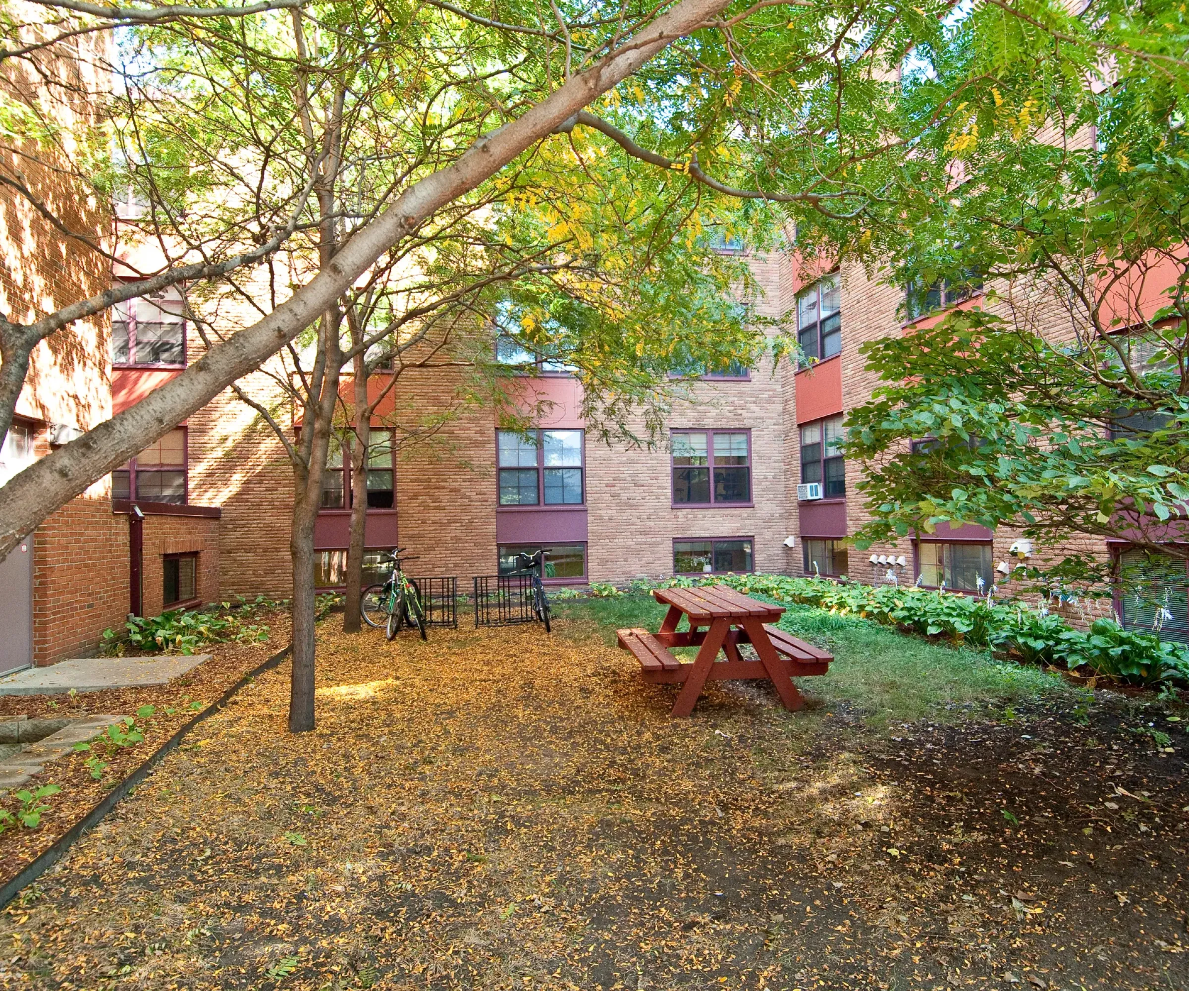 Aeon Affordable Homes and Community An affordable apartment home with a courtyard, trees, a red picnic table, fallen leaves, and bicycles against a brick building.