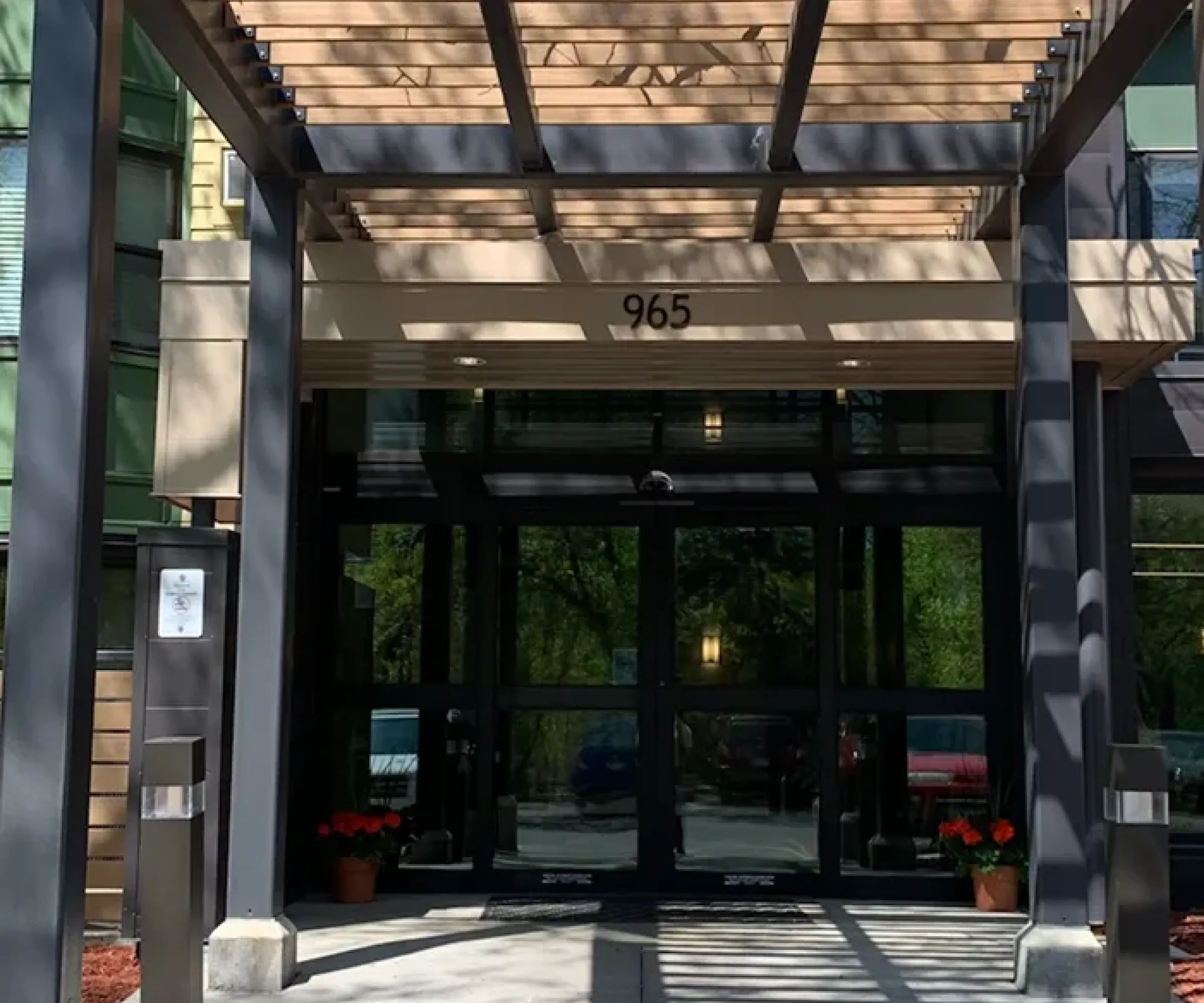 Aeon Affordable Homes and Community Entrance of affordable apartment homes with a wooden pergola and glass doors, casting patterned shadows on the walkway.