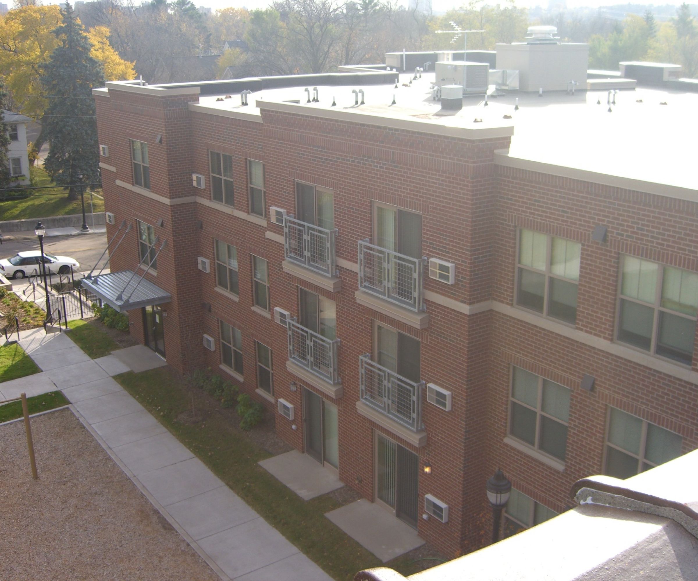 Aeon Affordable Homes and Community Affordable apartment homes in a brick building with balconies, visible air conditioning units, and a surrounding lawn with a playground area.