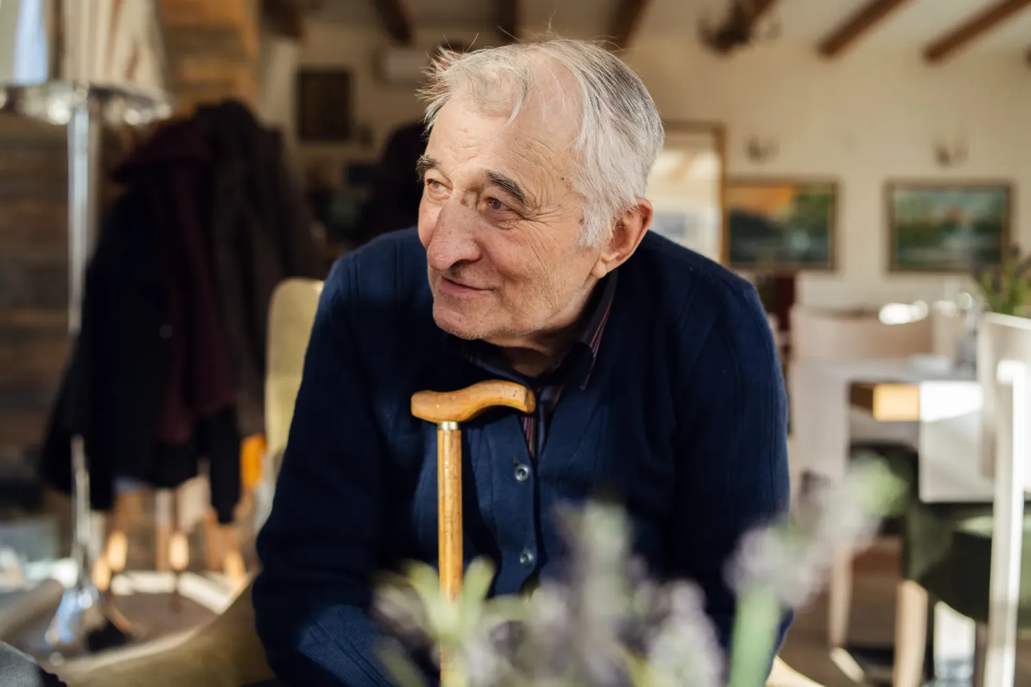 Aeon Affordable Homes with an elderly man in a cozy room, smiling and holding a walking cane. Paintings and coat in the background.