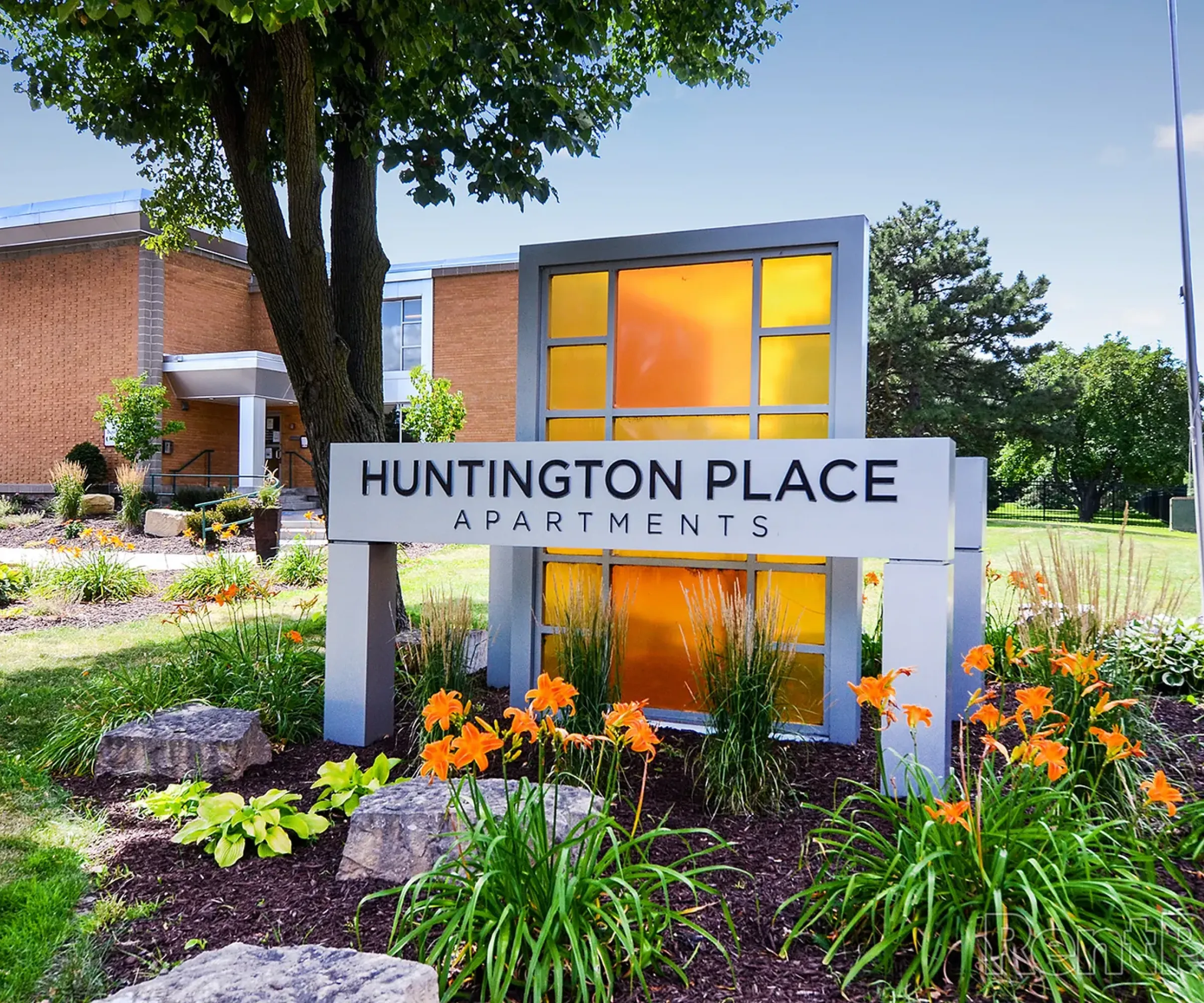 Aeon Affordable Homes Sign for Huntington Place Apartments, surrounded by flowers and greenery, with a modern building in the background.