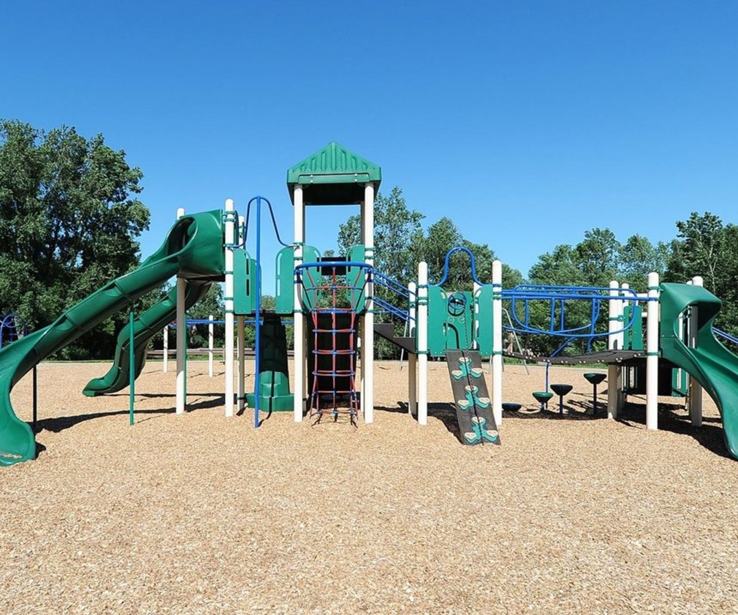 Aeon Affordable Homes and Community Playground with green slides and climbing structures, nestled among trees on a sunny day, near affordable apartment homes.