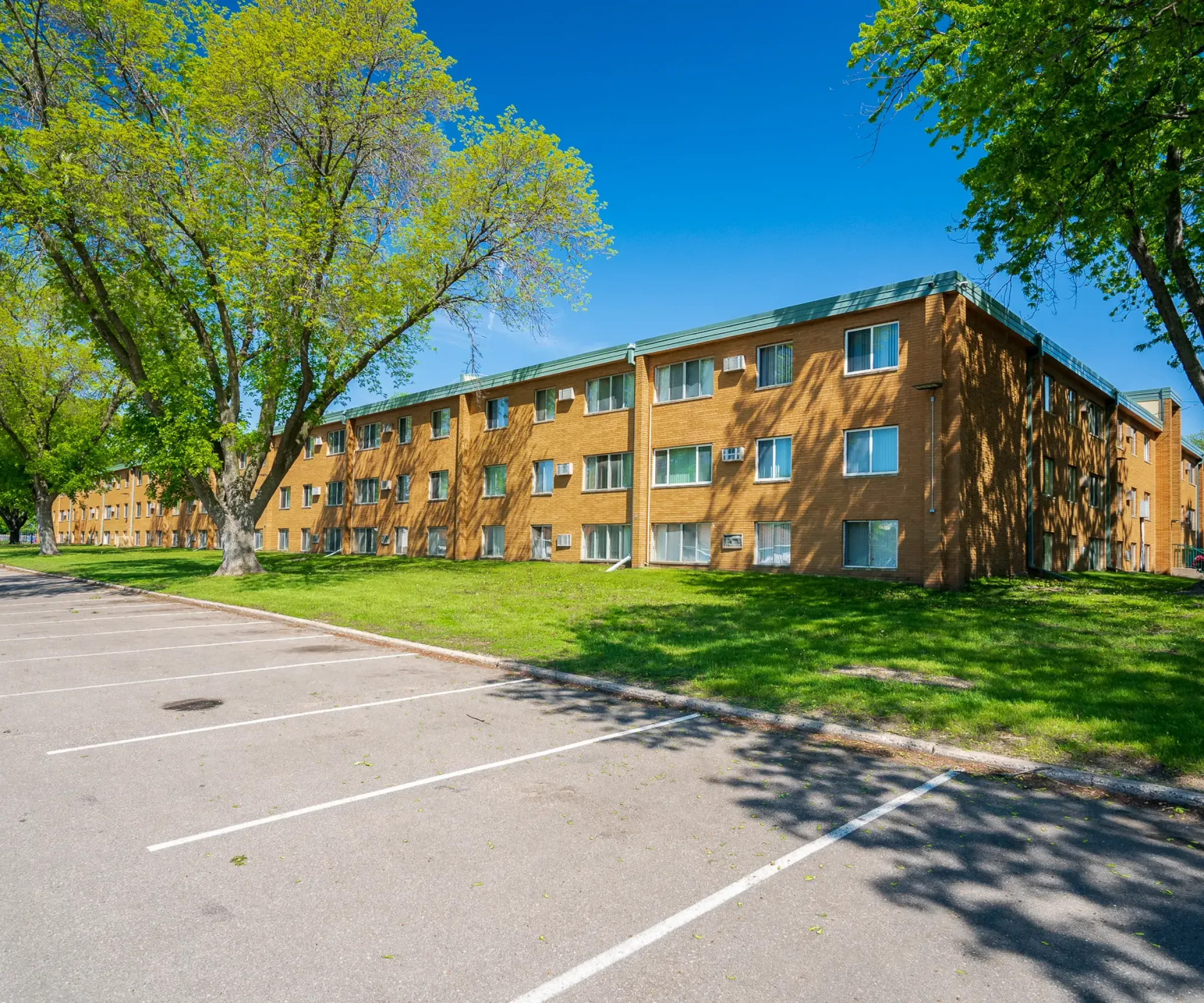 Aeon Affordable Homes A brick apartment building with a parking lot and trees under a clear blue sky.
