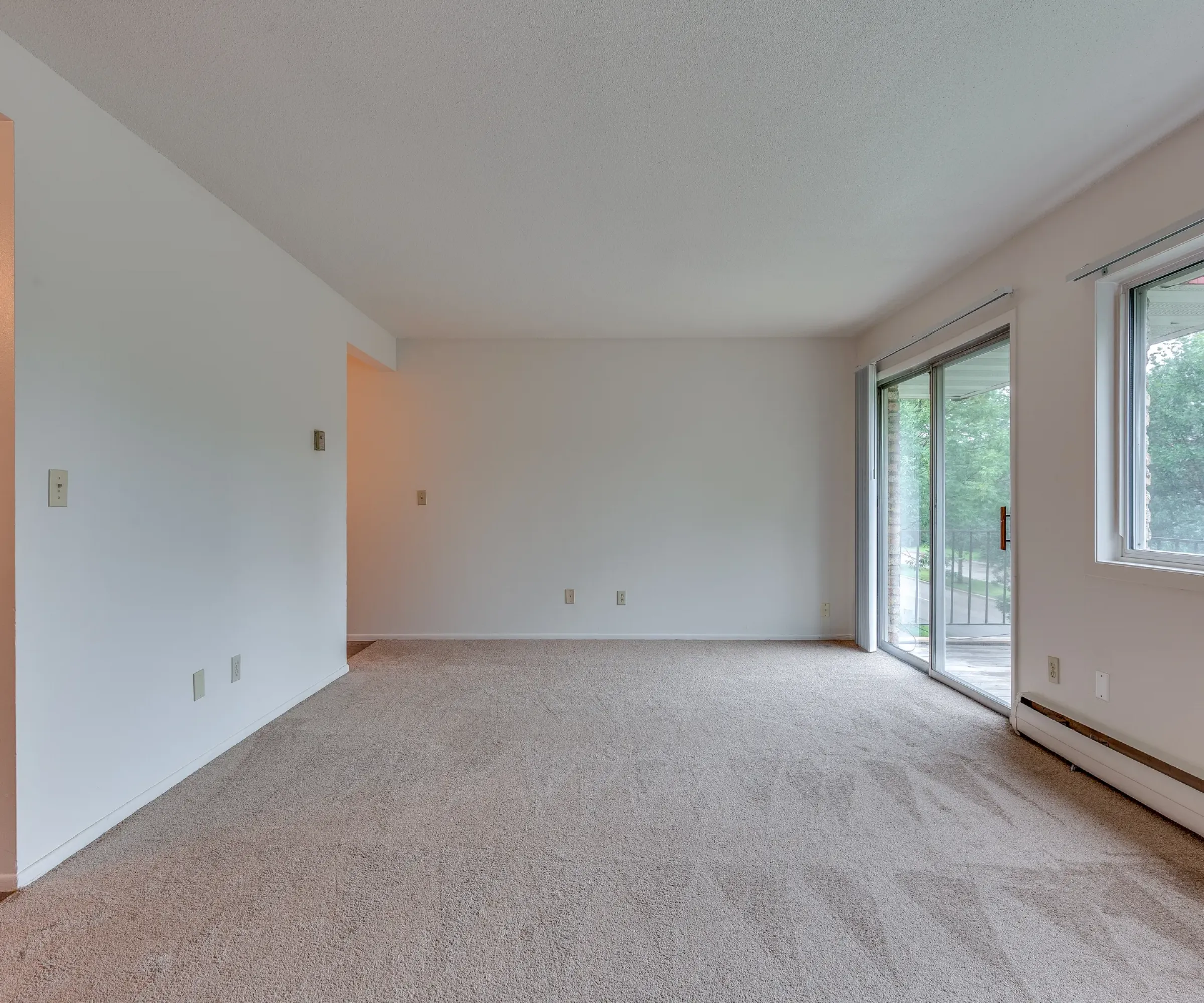 Aeon Affordable Homes and Community Affordable apartment homes featuring an empty carpeted room, large windows, and a sliding door leading to a patio near the partially visible kitchen.